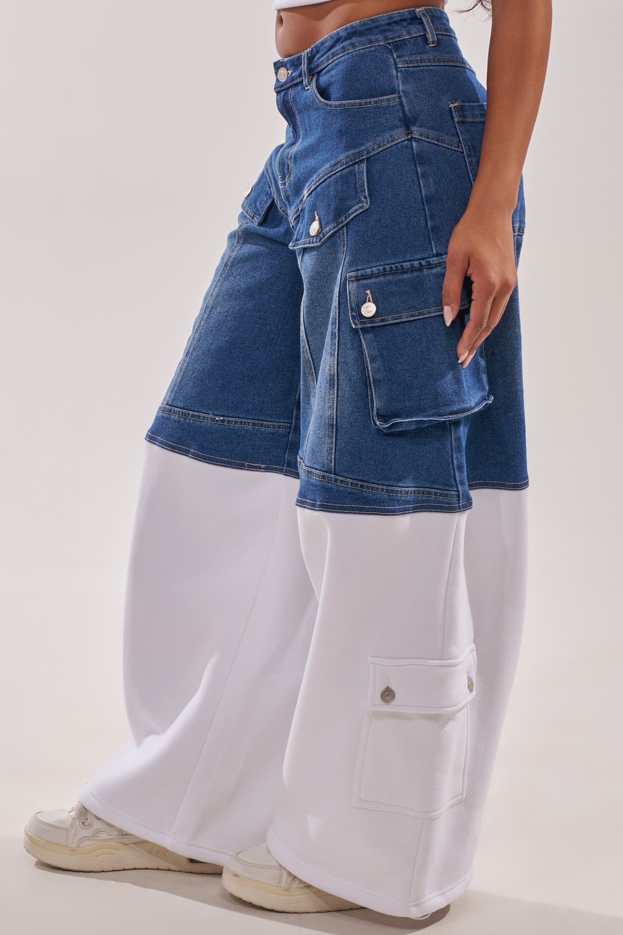 Someone wears the THIS IS EVERYTHING YOU SEE RELAXED DENIM—wide-leg streetwear pants with large cargo pockets on blue denim above and white fabric below—paired with white sneakers, against a plain light background. 3