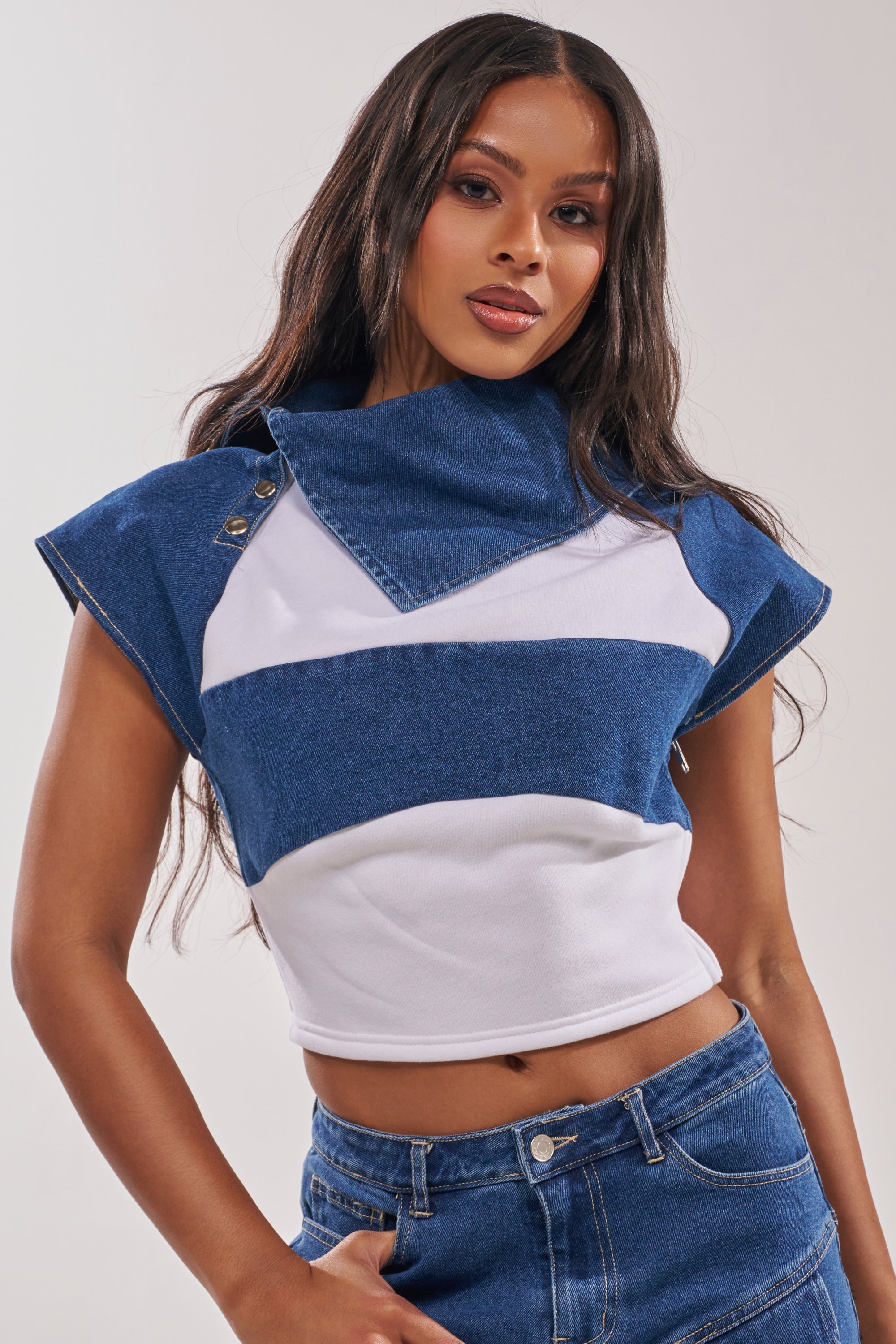 A woman poses against a neutral background wearing the THIS IS EVERYTHING YOU SEE DENIM BLOUSE—a cropped, fitted top with wide blue and white stripes and a mock-style oversized collar—paired with blue jeans. 0