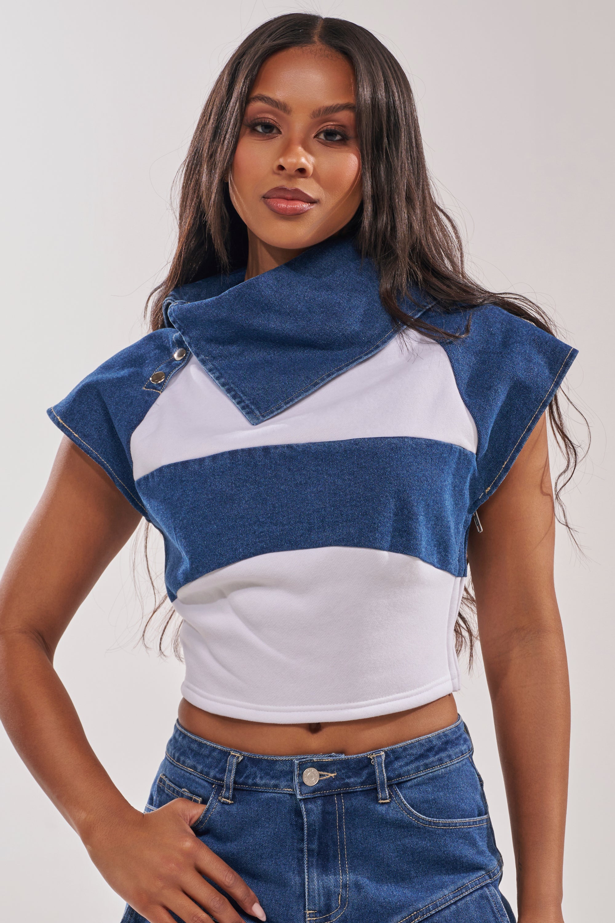 A woman wears the THIS IS EVERYTHING YOU SEE DENIM BLOUSE, a cropped fitted top with blue and white color-block design and an oversized asymmetrical collar, paired with blue denim jeans against a plain background. 1