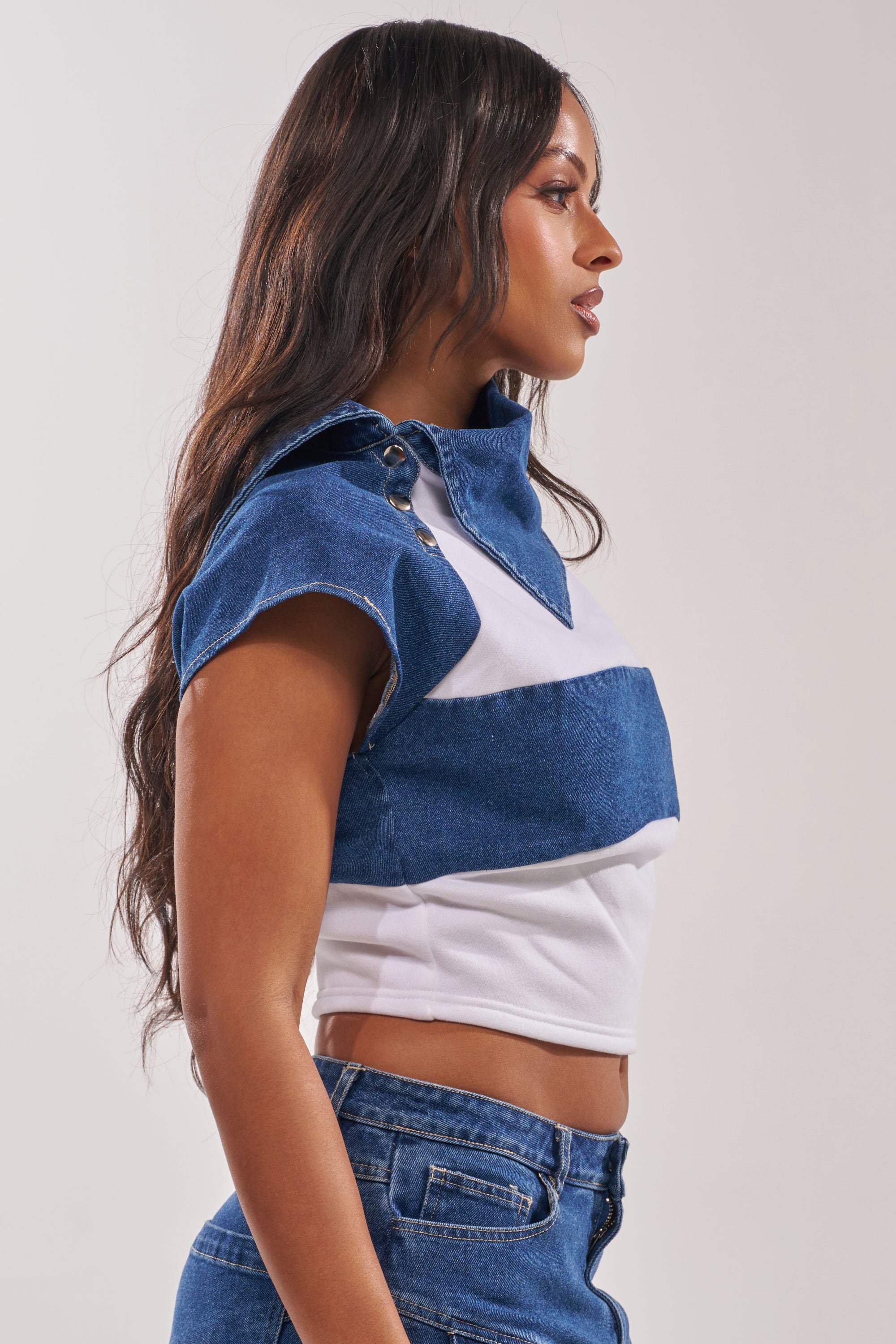 Woman with long dark hair wears the THIS IS EVERYTHING YOU SEE DENIM BLOUSE—a blue and white cropped fitted top with metallic collar fasteners—paired with matching blue denim jeans, standing in profile against a light background. 4