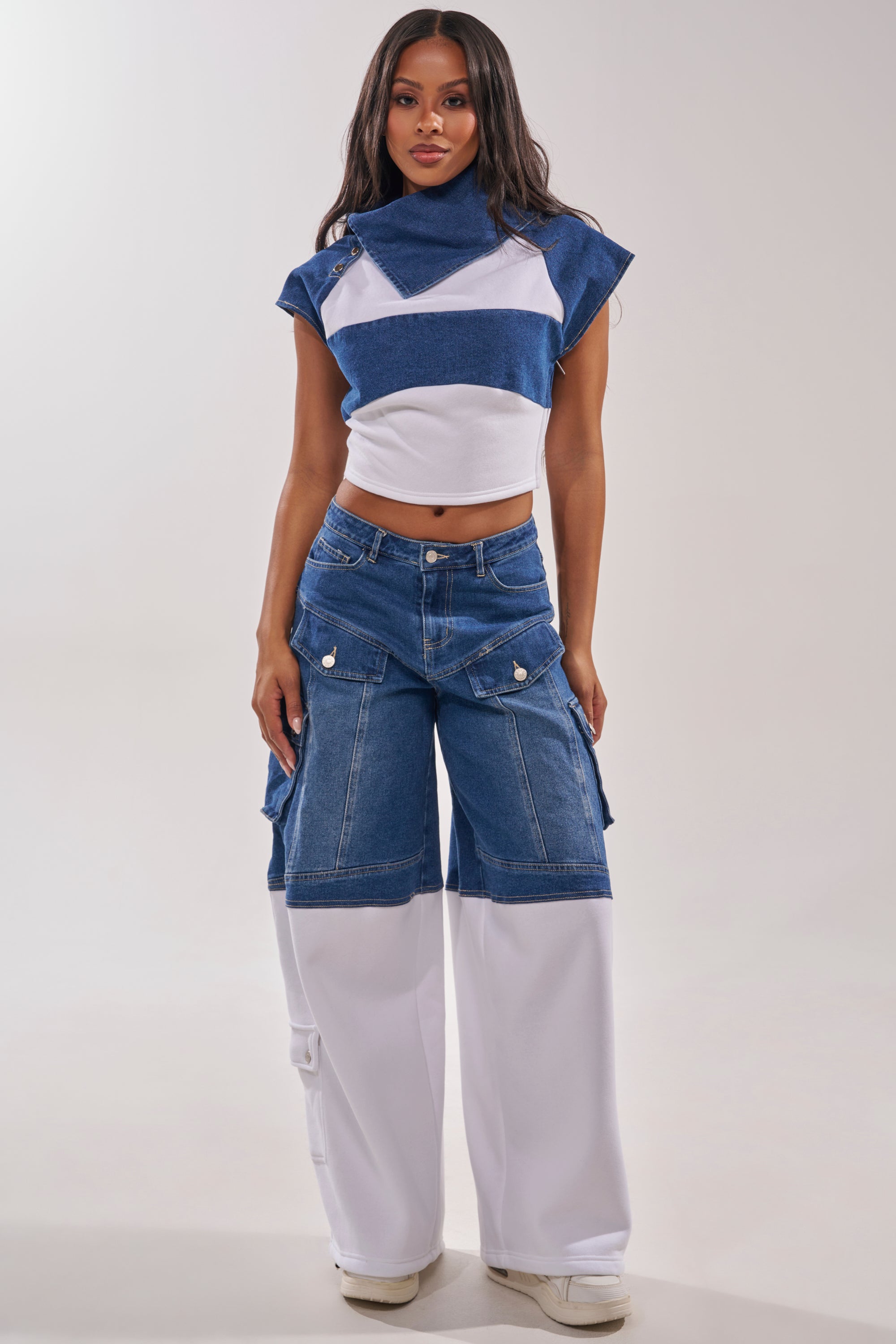 A woman faces forward wearing the THIS IS EVERYTHING YOU SEE DENIM BLOUSE, a blue and white two-tone fitted crop top with a high neck, paired with wide-leg jeans featuring denim and white panels. She has long hair and wears white sneakers against a plain background. 6
