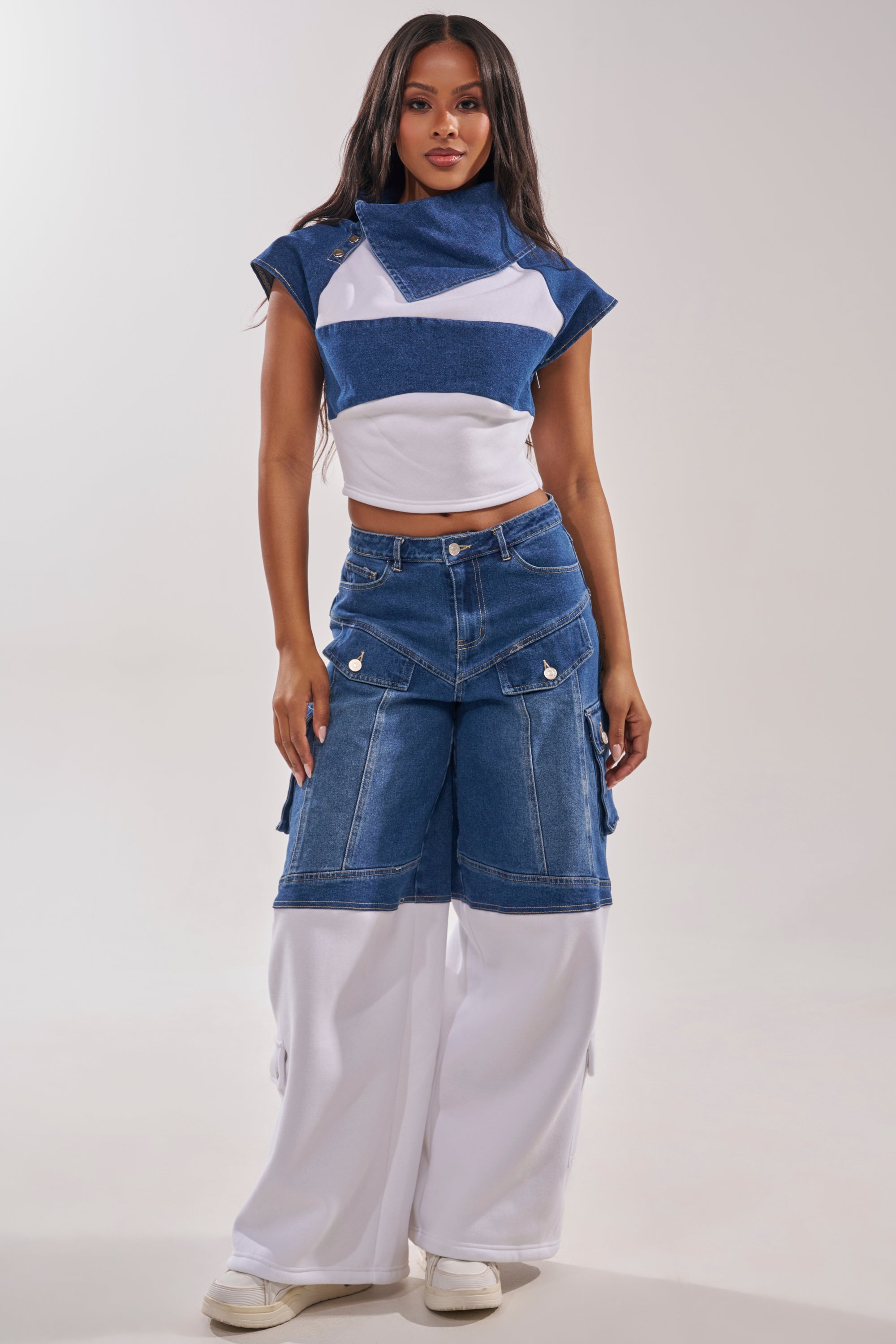 A woman with long hair and white sneakers poses against a plain light background wearing the THIS IS EVERYTHING YOU SEE DENIM BLOUSE, an asymmetrical cropped denim-and-cotton top, paired with wide-leg jeans featuring blue and white denim panels. 7