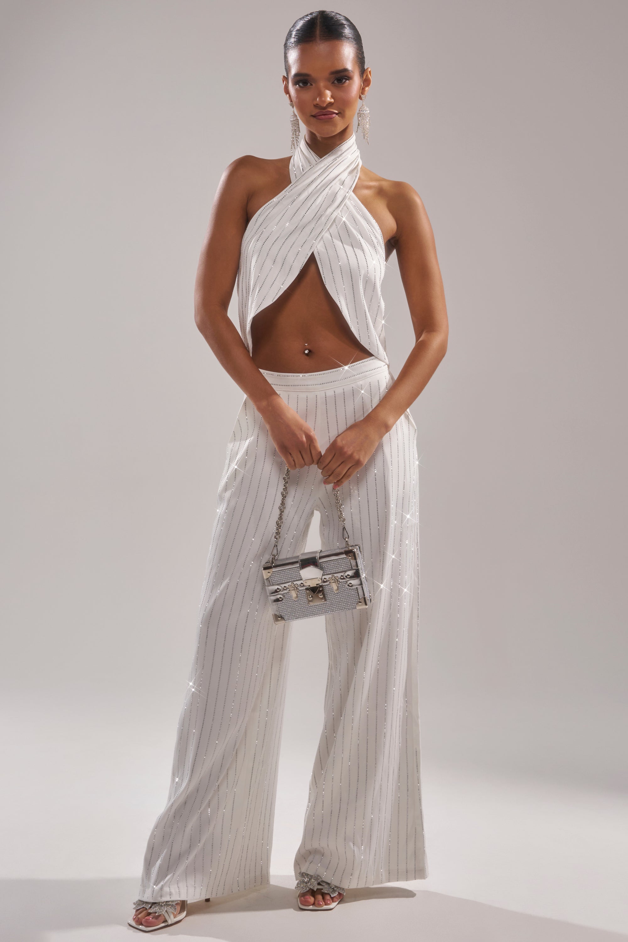 A woman poses confidently against a plain background, wearing the PRINCESS TREATMENT RHINESTONE WIDE LEG PANT with a white pinstriped halter top. She holds a metallic handbag, sporting minimal jewelry and a slicked-back hairstyle. 5