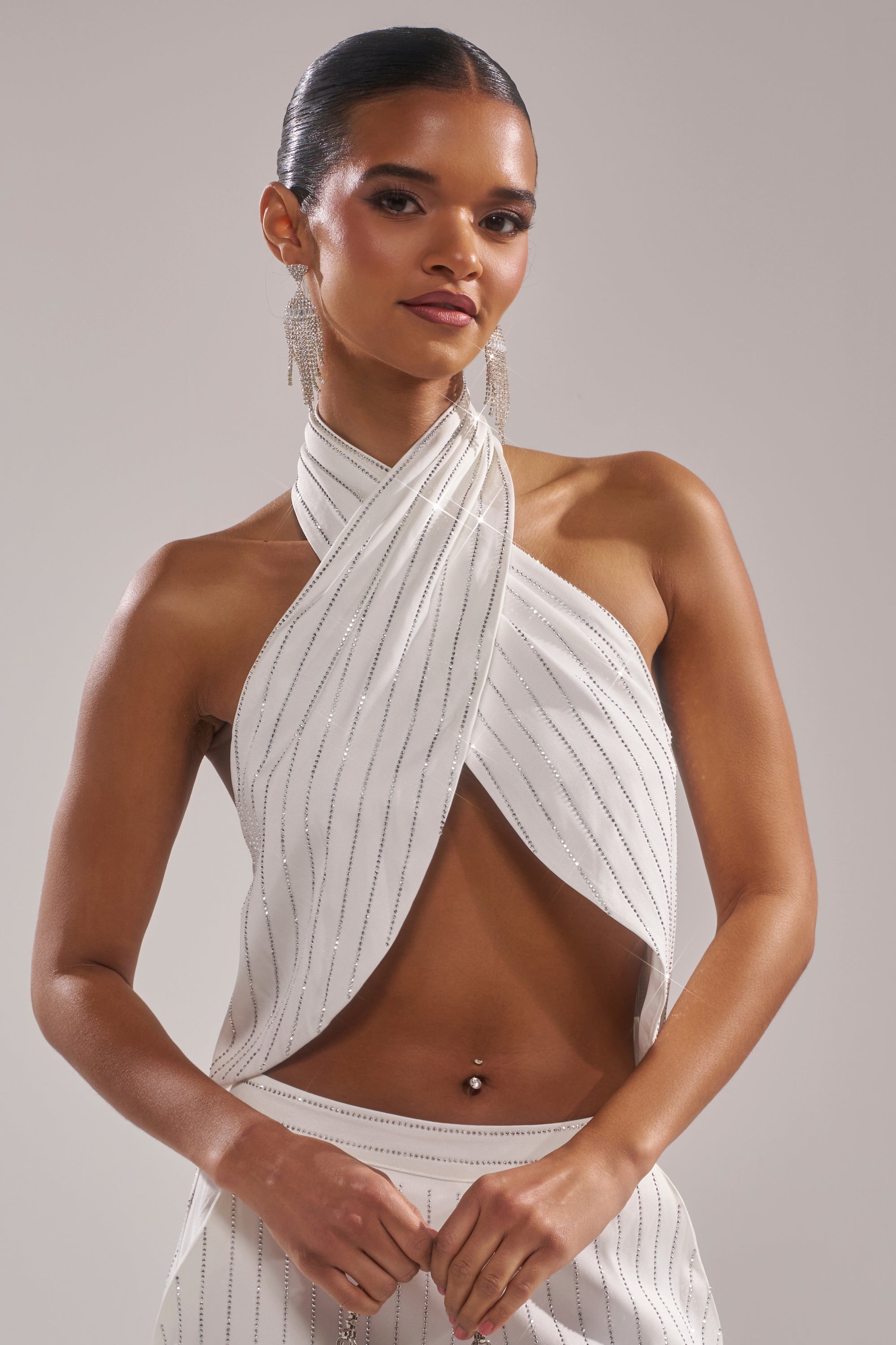 A woman with slicked-back hair styles the PRINCESS TREATMENT RHINESTONE HALTER TOP, featuring a crisscross design and matching skirt. She accessorizes with dangling earrings and a belly button piercing, posing against a neutral background. 0