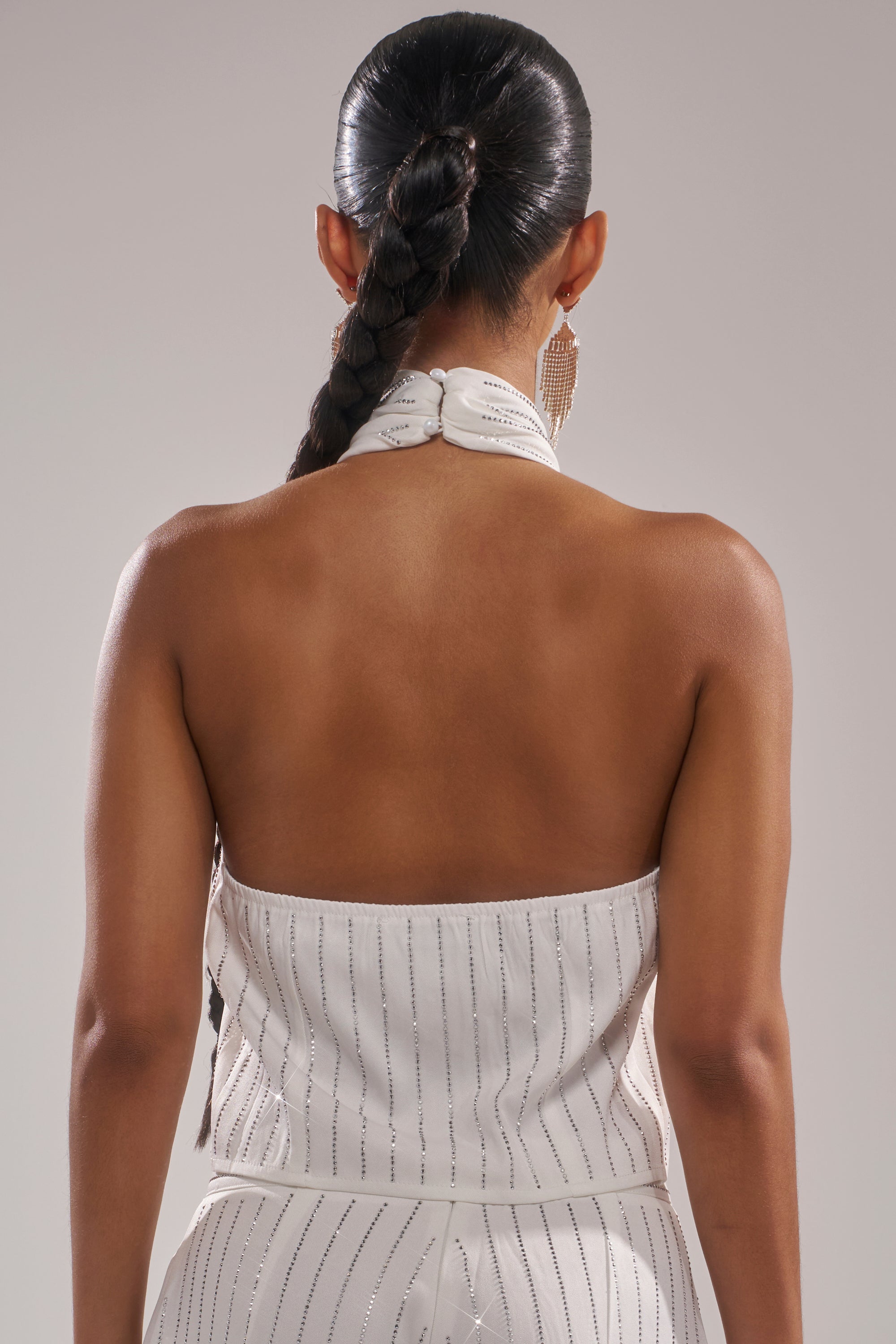 A woman with a long, braided ponytail is seen from the back, wearing the white, strapless PRINCESS TREATMENT RHINESTONE HALTER TOP and large dangling earrings. 3