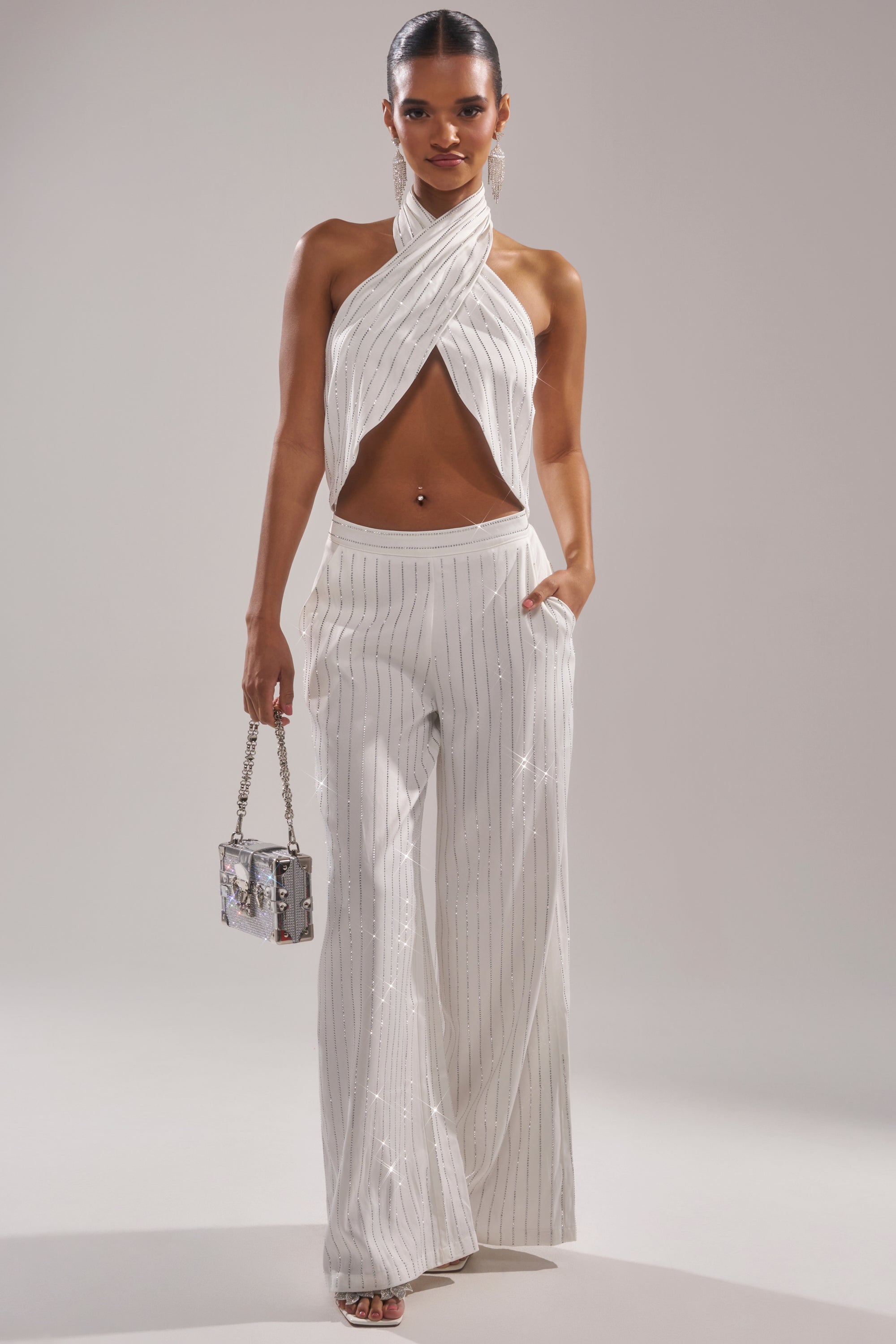A woman stands confidently in the PRINCESS TREATMENT RHINESTONE HALTER TOP, a white sleeveless halter with a crisscross front and rhinestone details, paired with matching wide-leg pants, a silver handbag, and open-toe heels for ultimate Princess Treatment style. 4