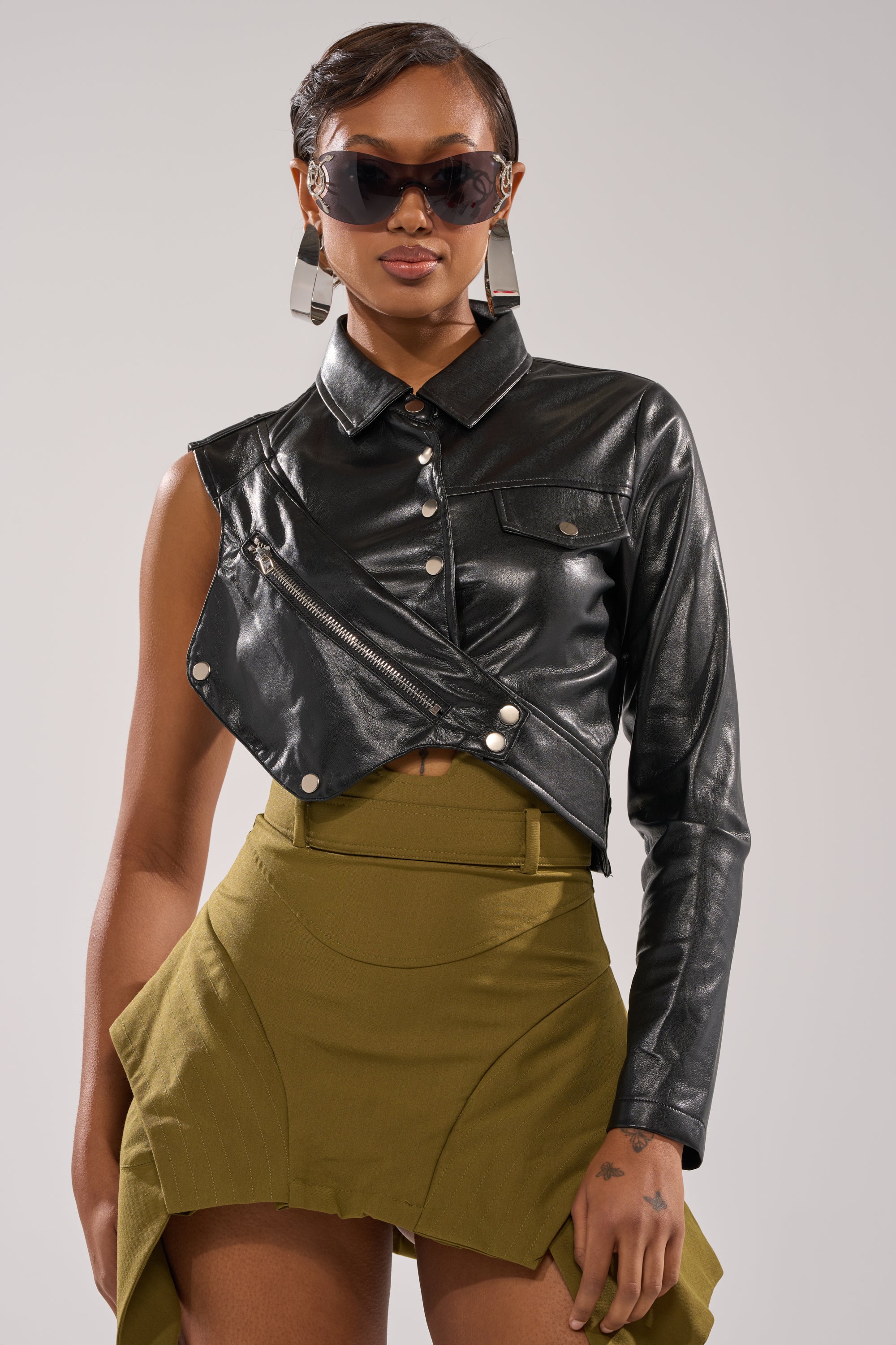 A woman confidently styles the ALL FOR ME FAUX LEATHER BLOUSE—an asymmetrical, black, one-sleeve biker top—with large sunglasses, geometric earrings, and a high-waisted olive green skirt against a plain background. 0