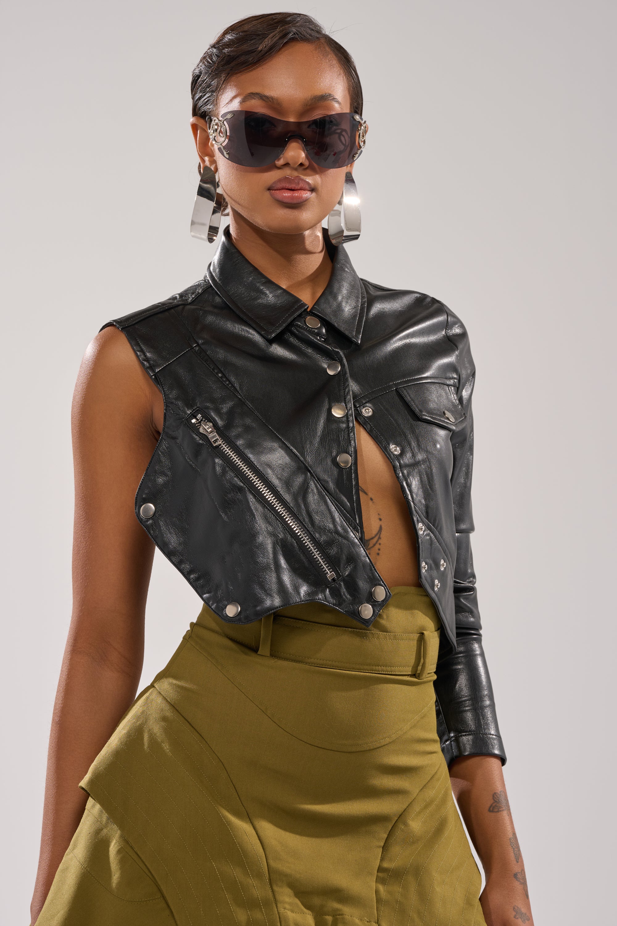 A stylish woman in the ALL FOR ME FAUX LEATHER BLOUSE, large sunglasses, chunky silver earrings, and an olive green high-waisted skirt poses confidently against a plain background. 1