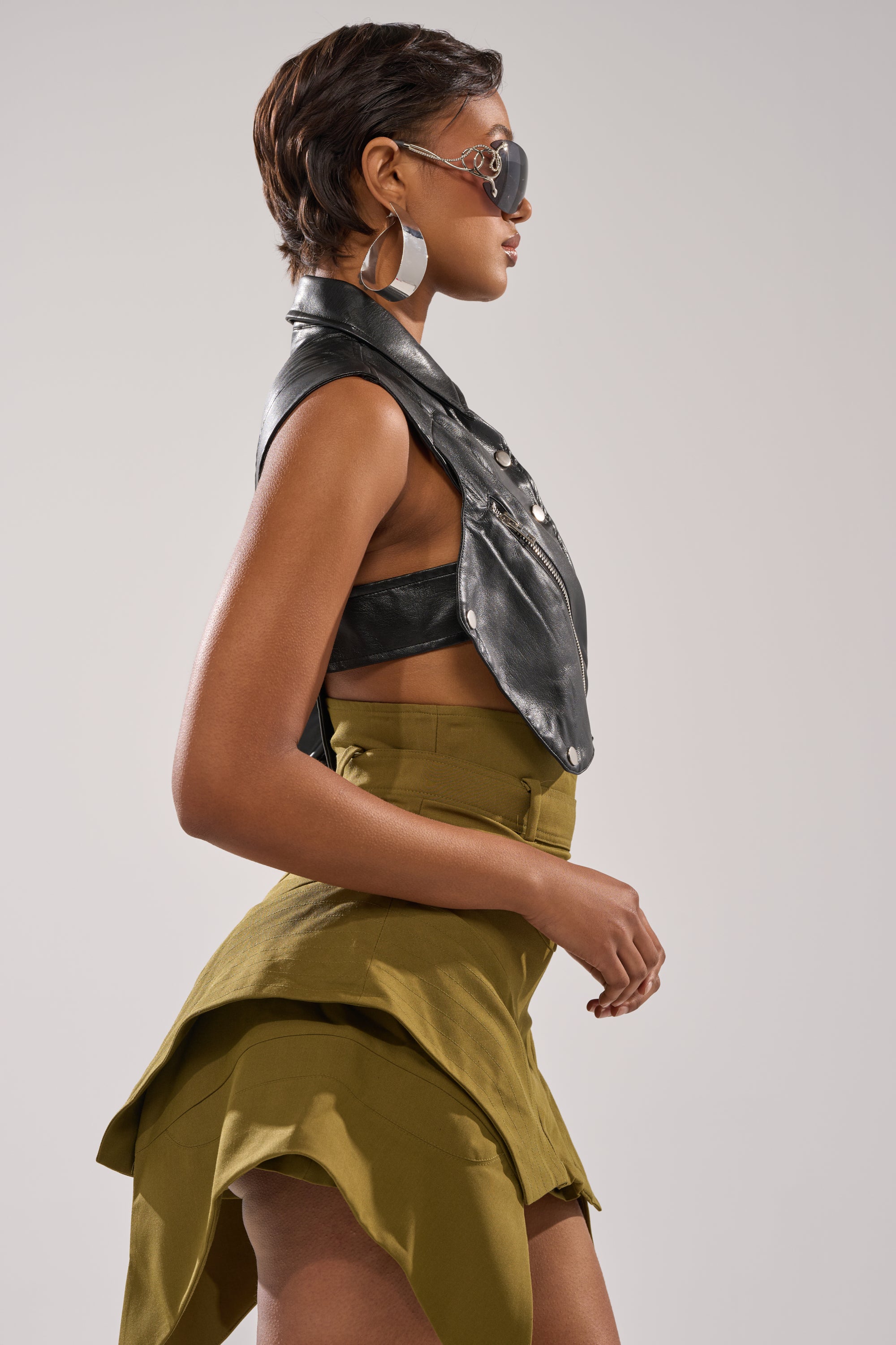 A woman in profile models the ALL FOR ME FAUX LEATHER BLOUSE in a cropped biker style with large hoop earrings, round sunglasses, and an olive green asymmetrical skirt against a plain light background. 3