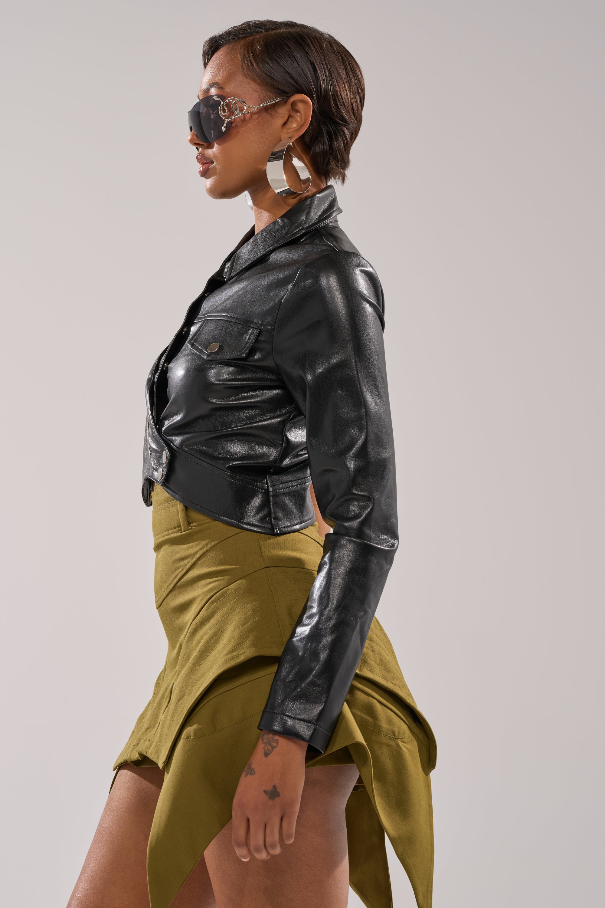 Wearing the ALL FOR ME FAUX LEATHER BLOUSE in black, a biker-style cropped jacket, paired with an olive green asymmetric skirt, large hoop earrings, and oversized sunglasses, posed sideways against a plain background. 4