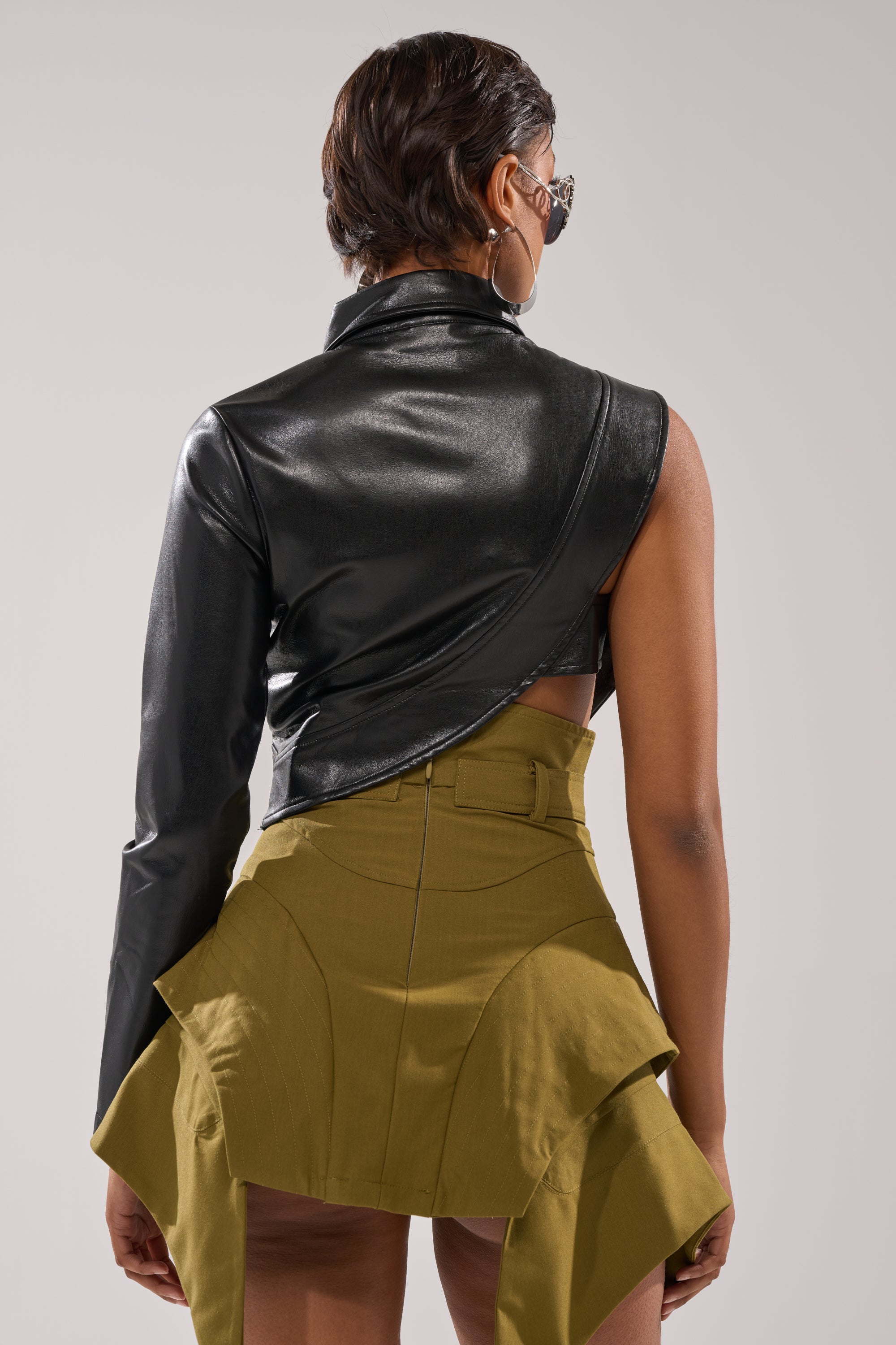 A person with short hair and hoop earrings wears the ALL FOR ME FAUX LEATHER BLOUSE, a cropped black biker-style top with one sleeve, paired with an olive green skirt with dramatic angled panels, shown from the back against a plain background. 5