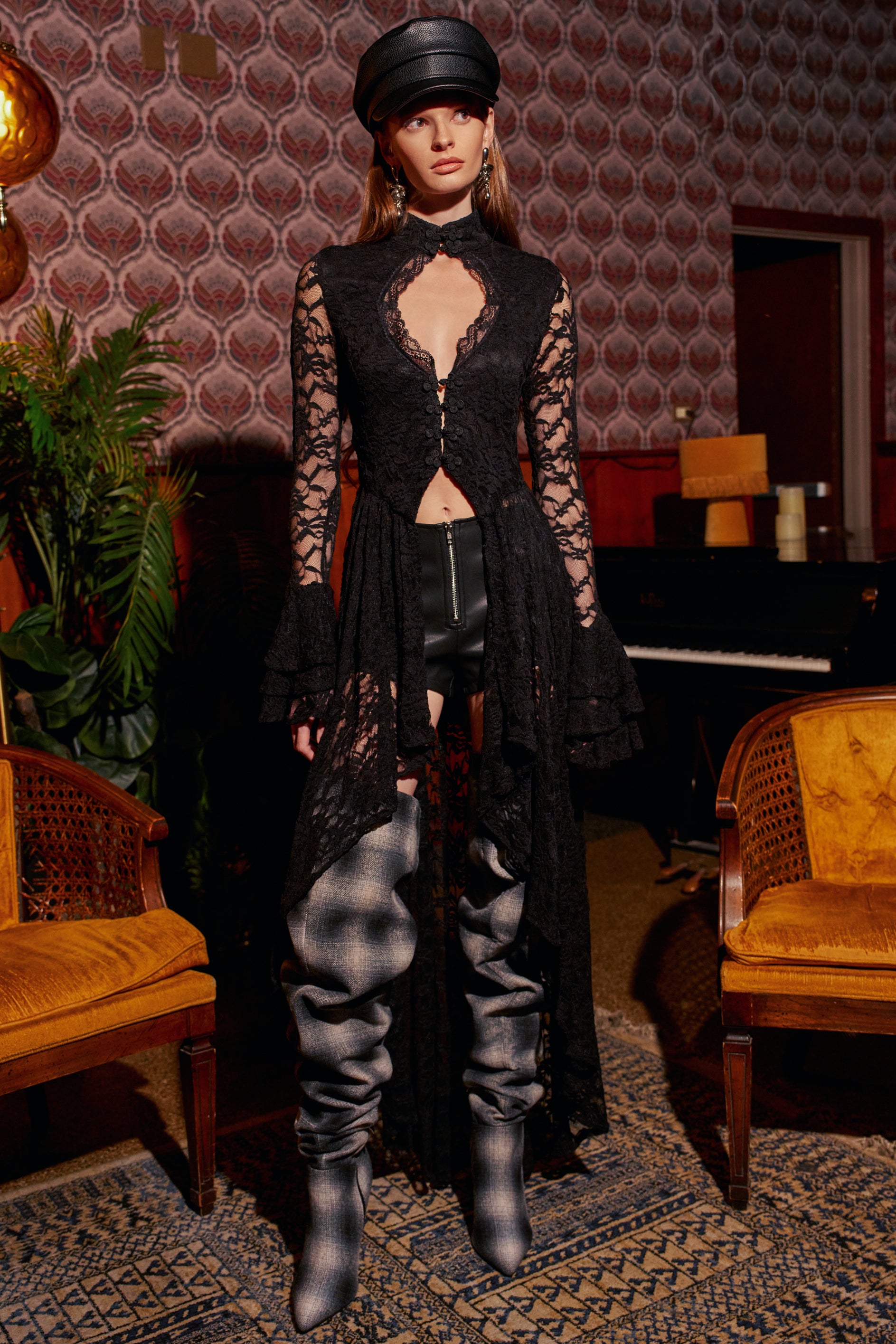 A model wears a BELINHA LACE BLOUSE IN BLACK, featuring a front cutout and a high-low hem, paired with black shorts and gray plaid thigh-high boots.