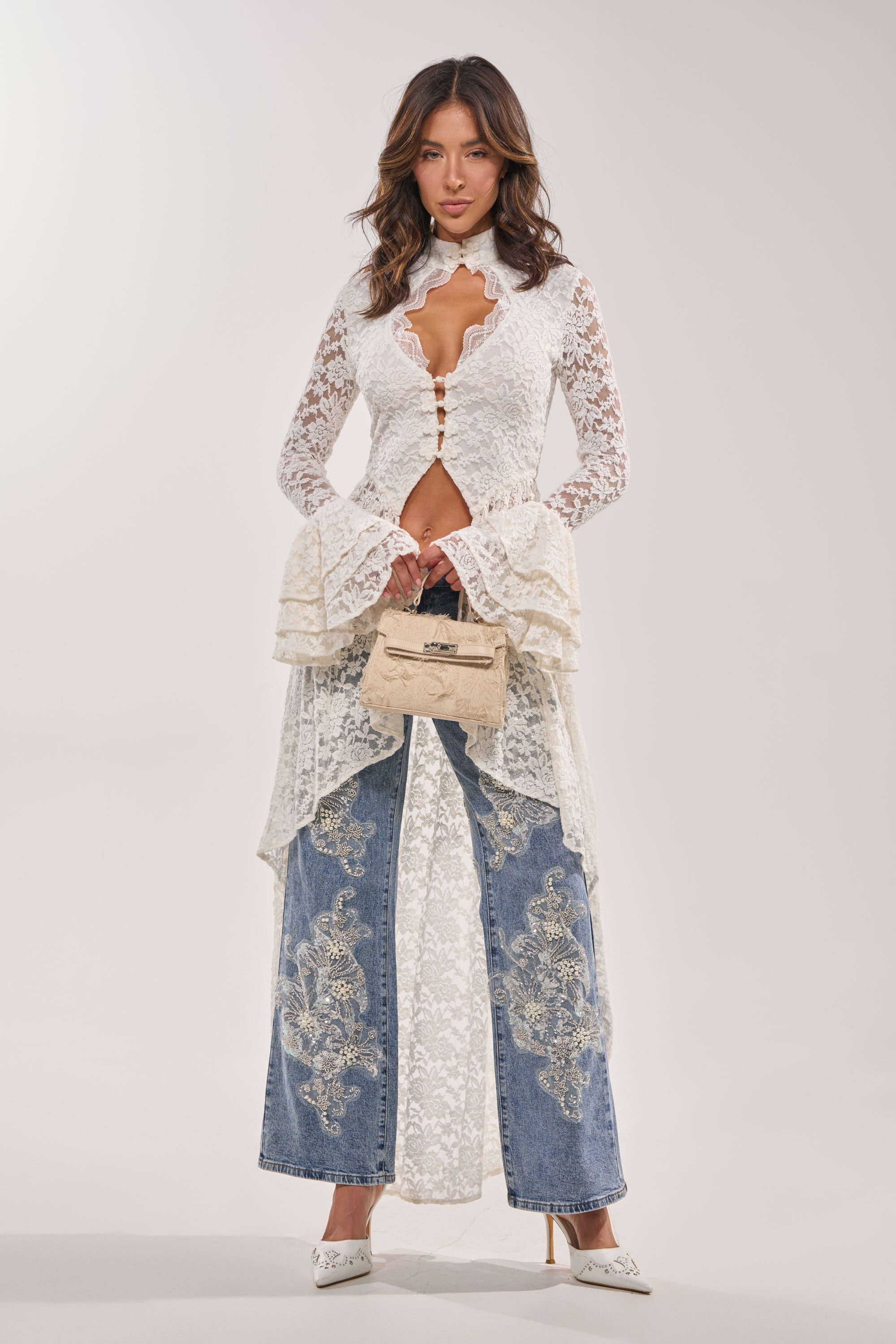 A woman stands against a plain light background wearing the BELINHA LACE BLOUSE, featuring ivory lace, flared sleeves, and an asymmetrical high-low hem, styled with embroidered blue jeans, a beige handbag, and white heels.