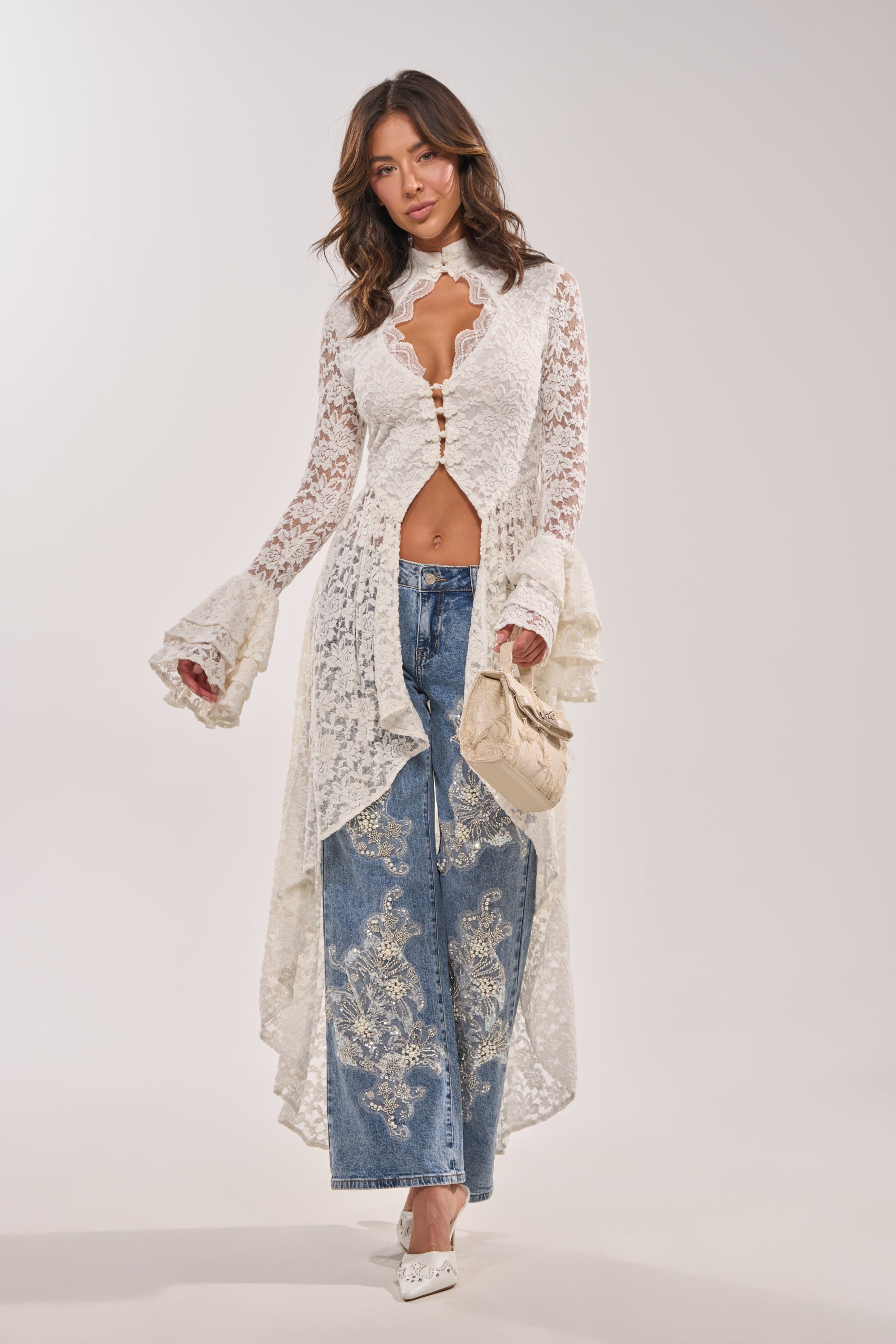 A woman models the BELINHA LACE BLOUSE, an elegant ivory top with ruffled sleeves, paired with embroidered wide-leg blue jeans, a beige purse, and white shoes as she poses stylishly against a plain light background.