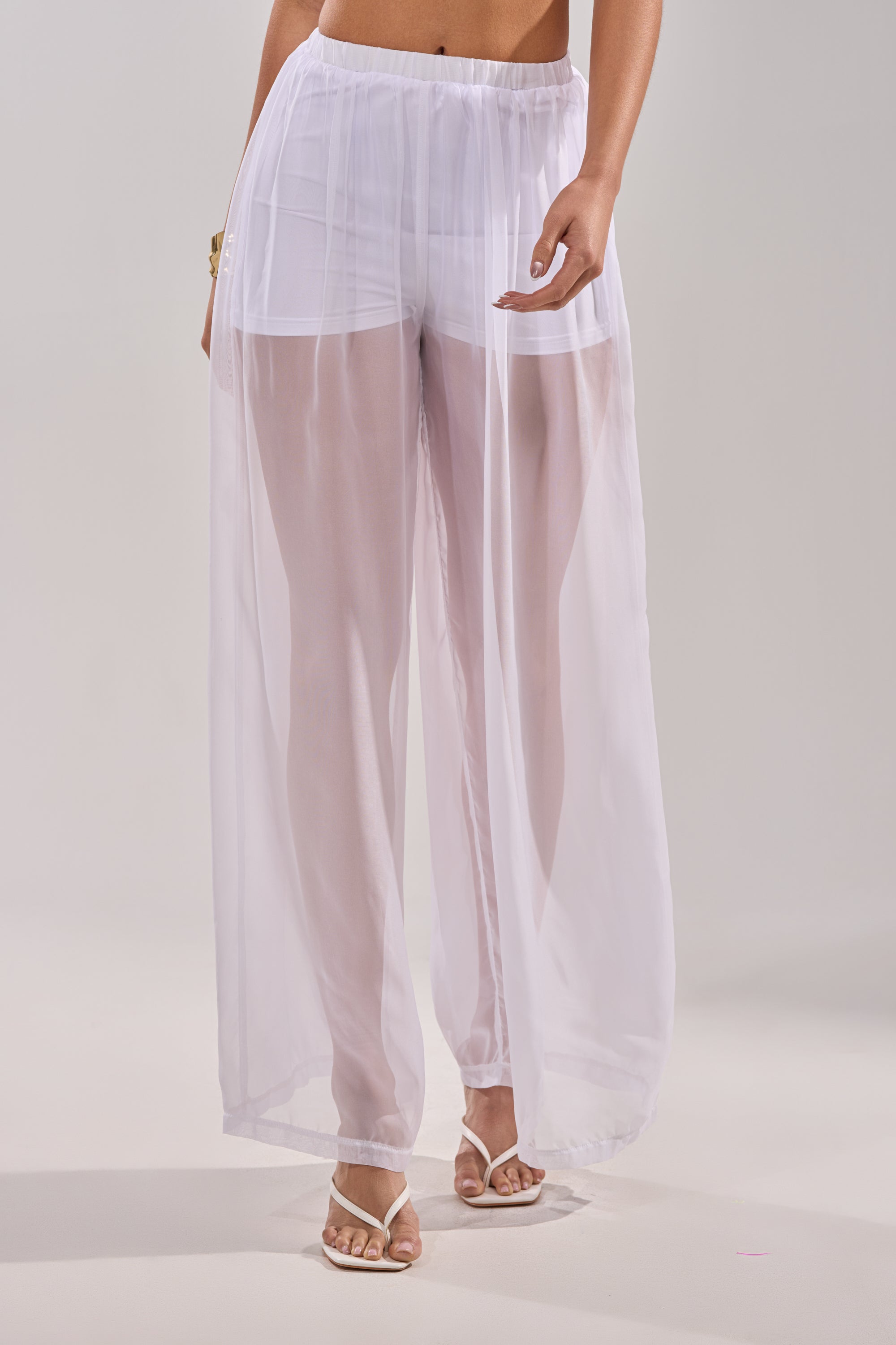 A woman models the DREAMY PALAZZO PANT—sheer wide-leg pants with built-in shorts—ideal for poolside layering, styled with strappy heeled sandals. The background is plain and neutral. 2