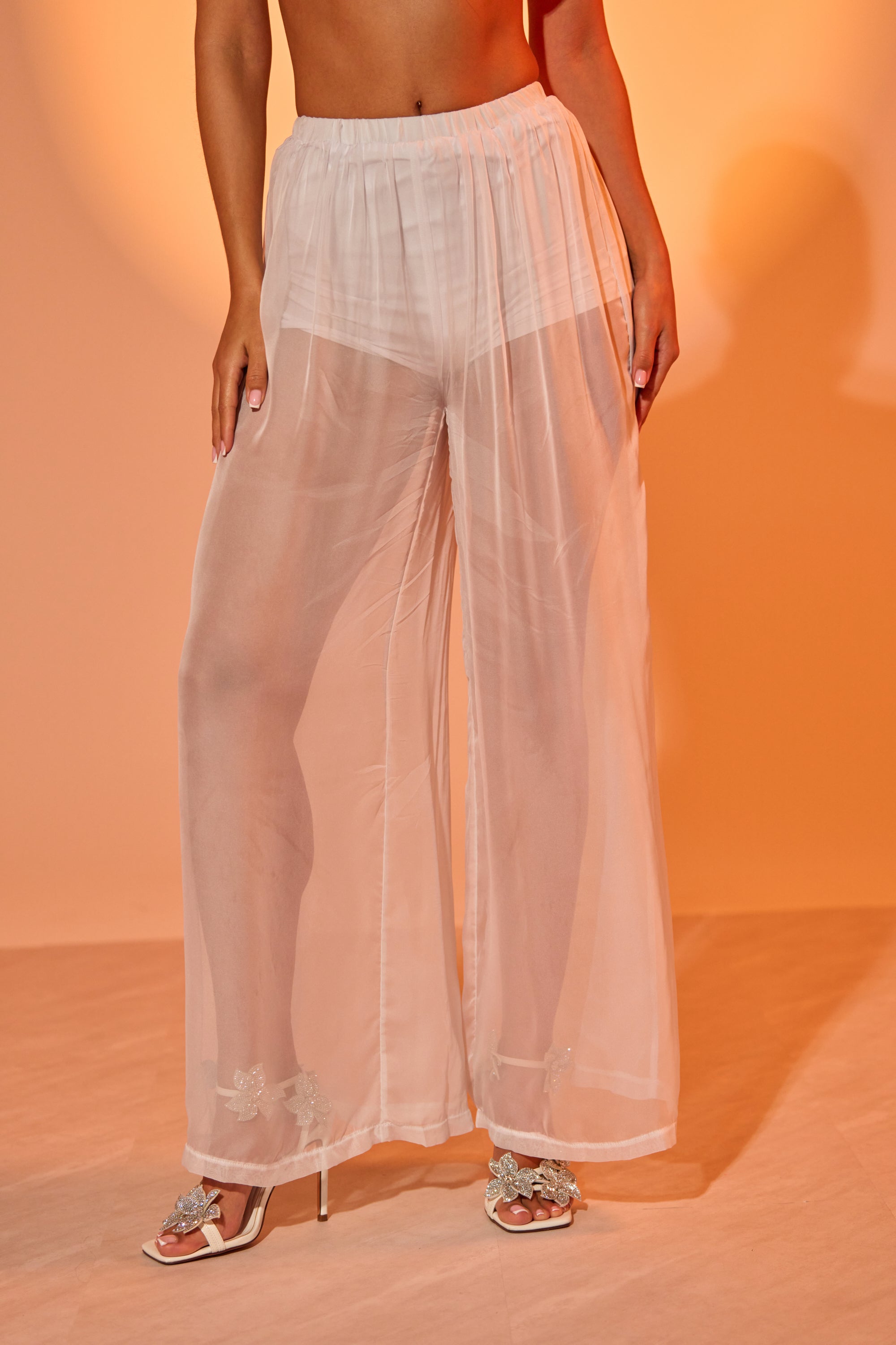 Wearing the DREAMY PALAZZO PANT—sheer, wide-leg mesh with built-in shorts—paired with floral-embellished heeled sandals against a peach backdrop, creating a chic, layered look ideal for poolside lounging. 1