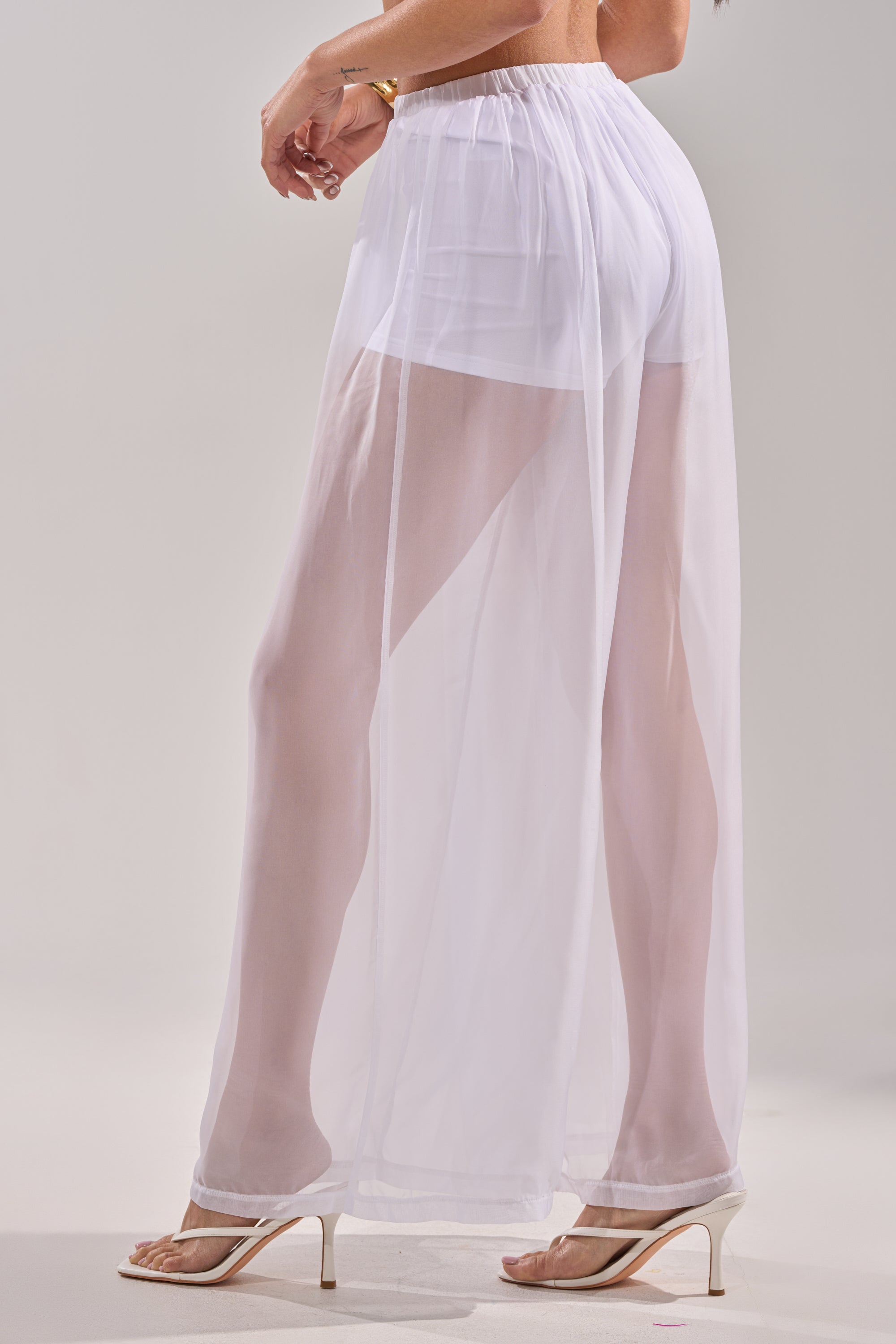 A person stands on a light surface, wearing the DREAMY PALAZZO PANT over white shorts with clear high-heeled sandals. This sheer wide-leg pant is perfect for chic poolside layering. Photo shows lower half only. 4