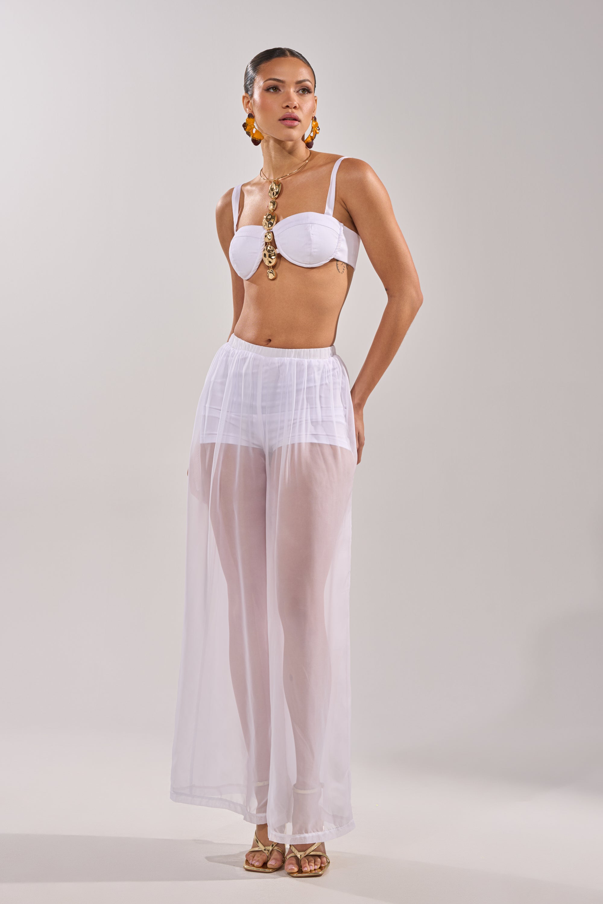 A woman poses in the DREAMY BRALETTE, a white underwire bralette with a sweetheart neckline, paired with sheer, high-waisted wide-leg pants, gold sandals, statement earrings, and a bold gold necklace against a plain light background. 6