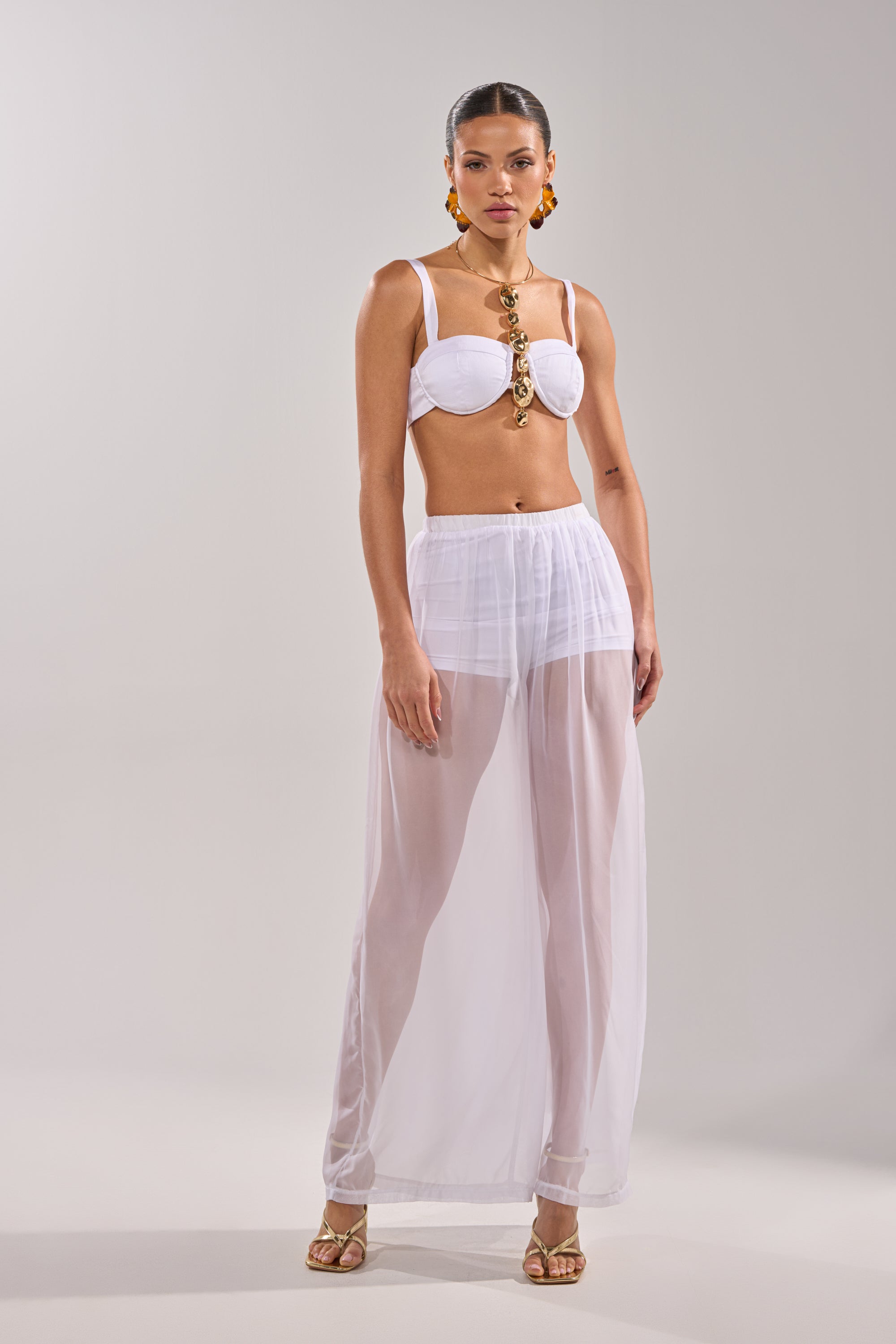 A woman poses against a neutral background wearing the DREAMY BRALETTE with a smocked back band, sheer white wide-leg pants, gold sandals, bold jewelry, and her hair pulled back. 7