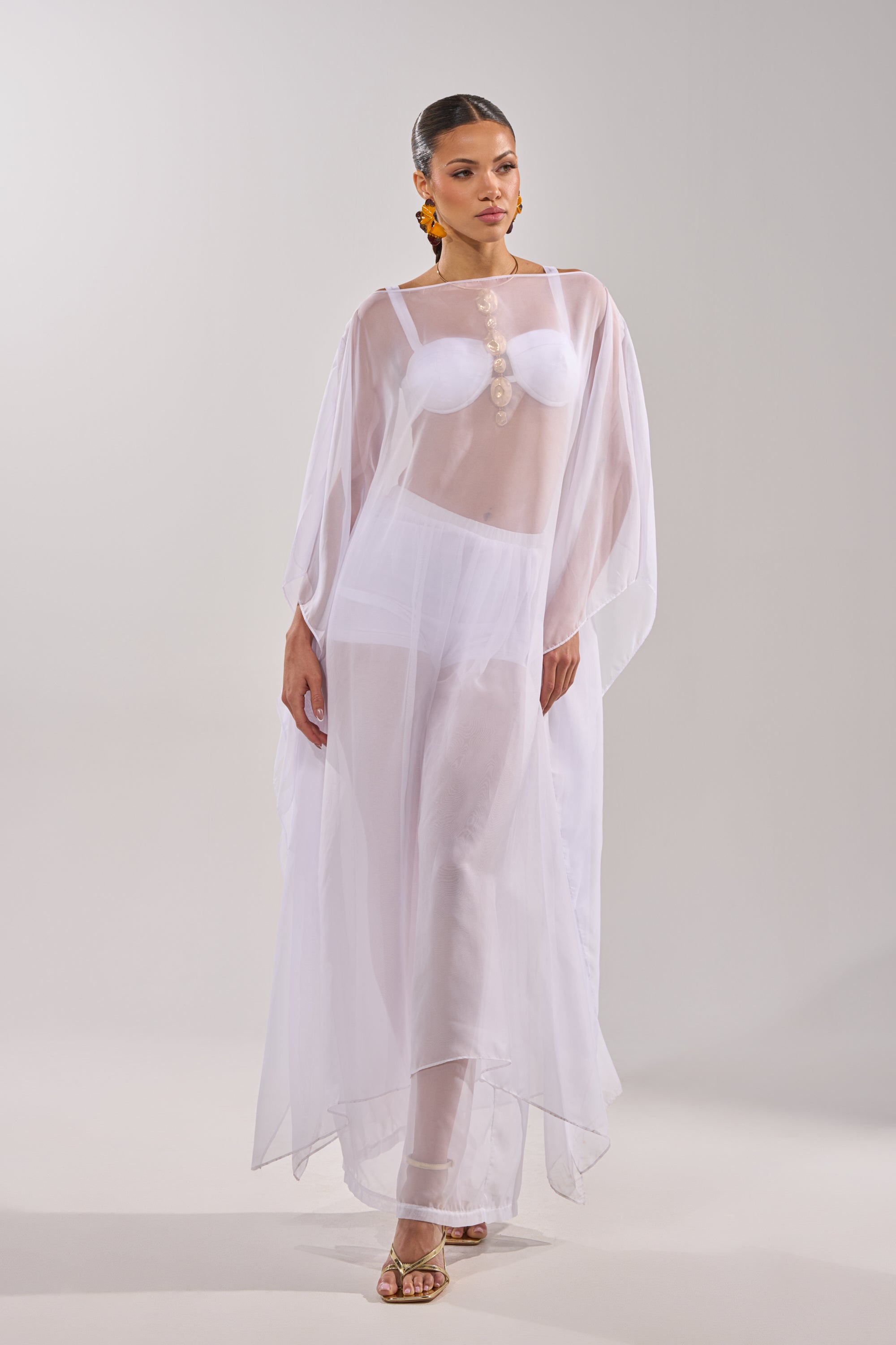 A woman poses against a light gray background in the DREAMY BRALETTE and briefs under a sheer, flowing white dress with a sweetheart neckline. She finishes the look with orange statement earrings and heeled sandals. 8