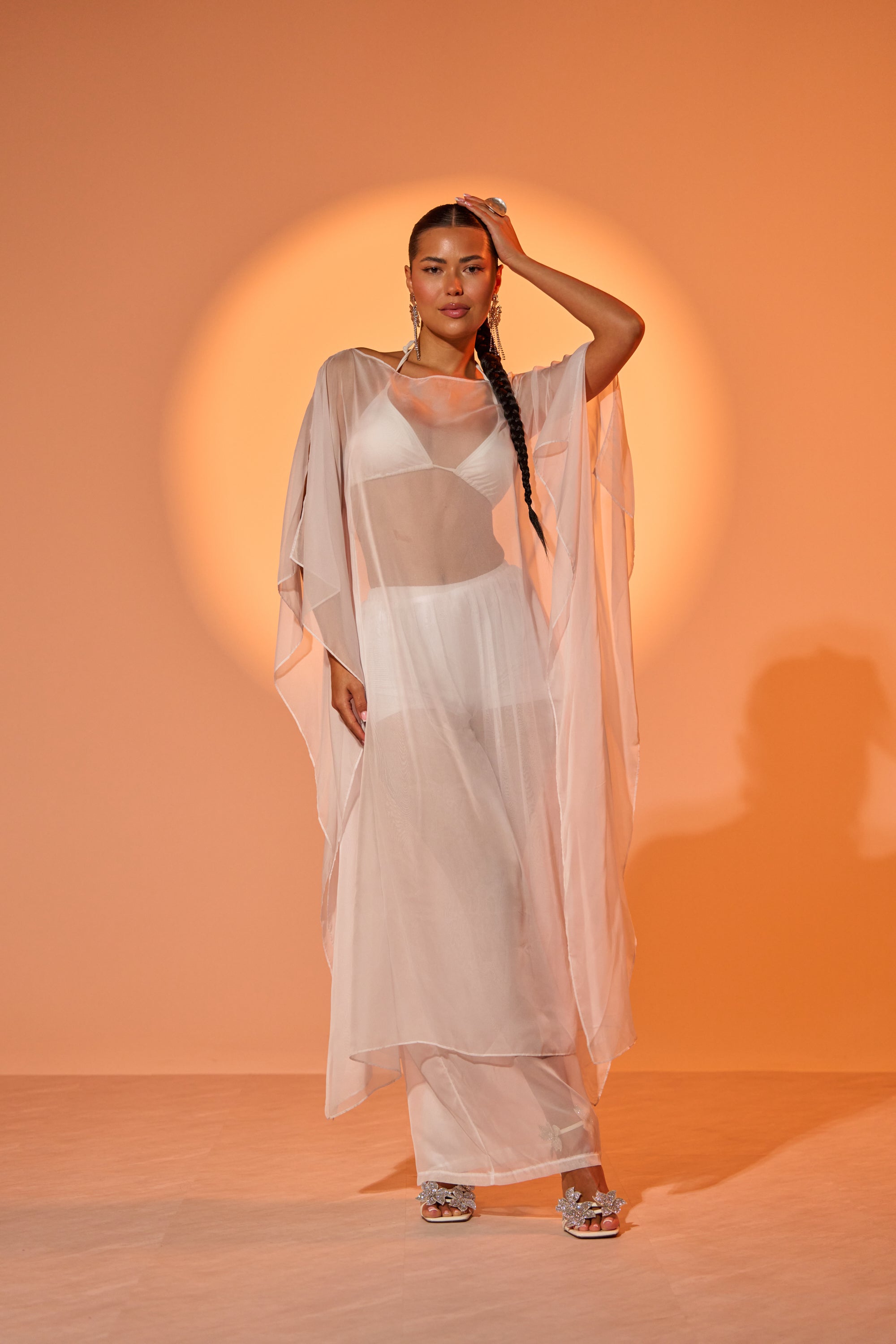 A woman with a long braid poses confidently in the DREAMY SHEER BLOUSE, layered over a bralette and skirt with a flowing white dress, standing before a warm orange background illuminated by a circular light. 0