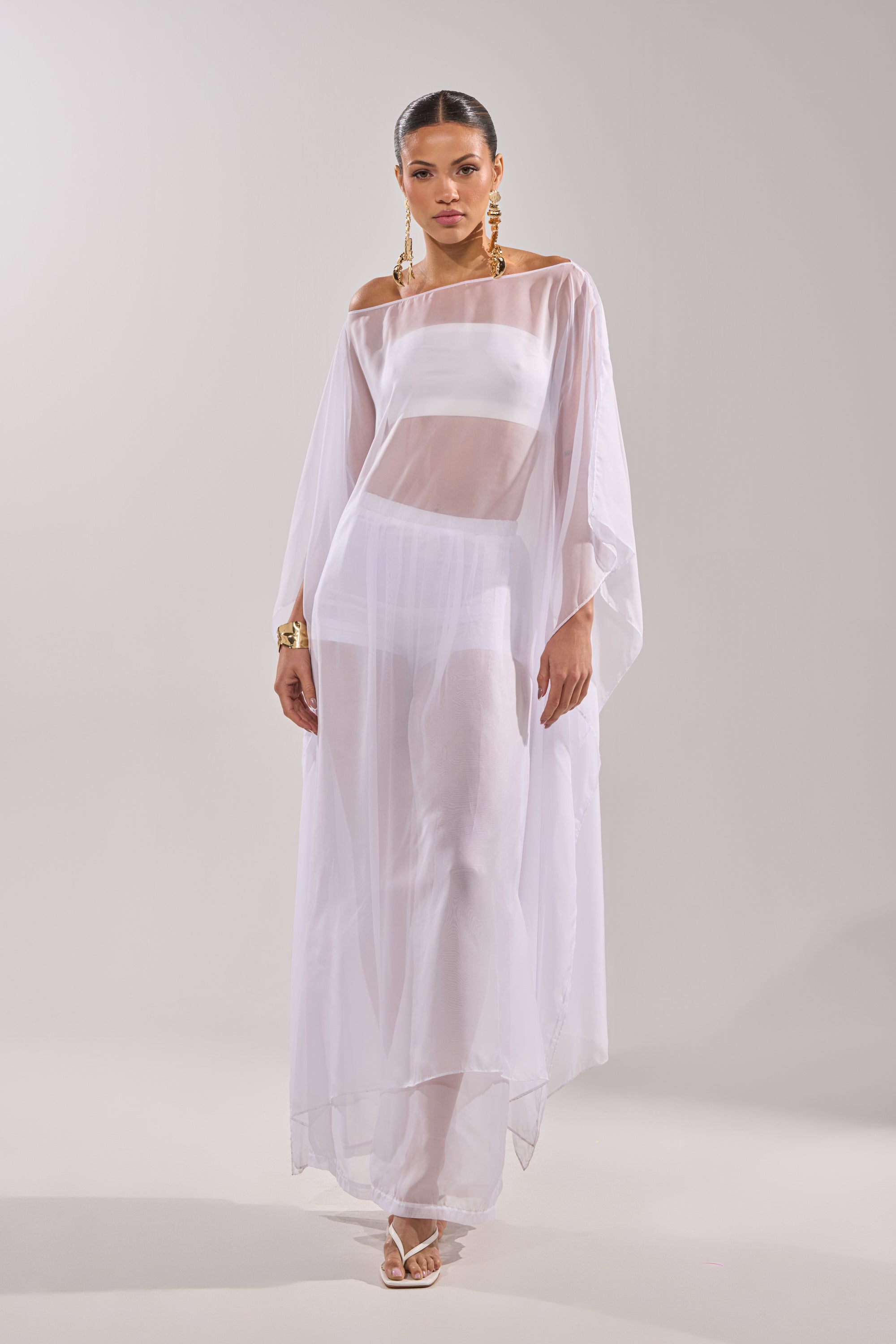 A woman models the DREAMY SHEER BLOUSE, styled as a flowing white off-the-shoulder dress. She pairs it with gold earrings, a gold bracelet, and white heeled sandals against a plain light background. 3
