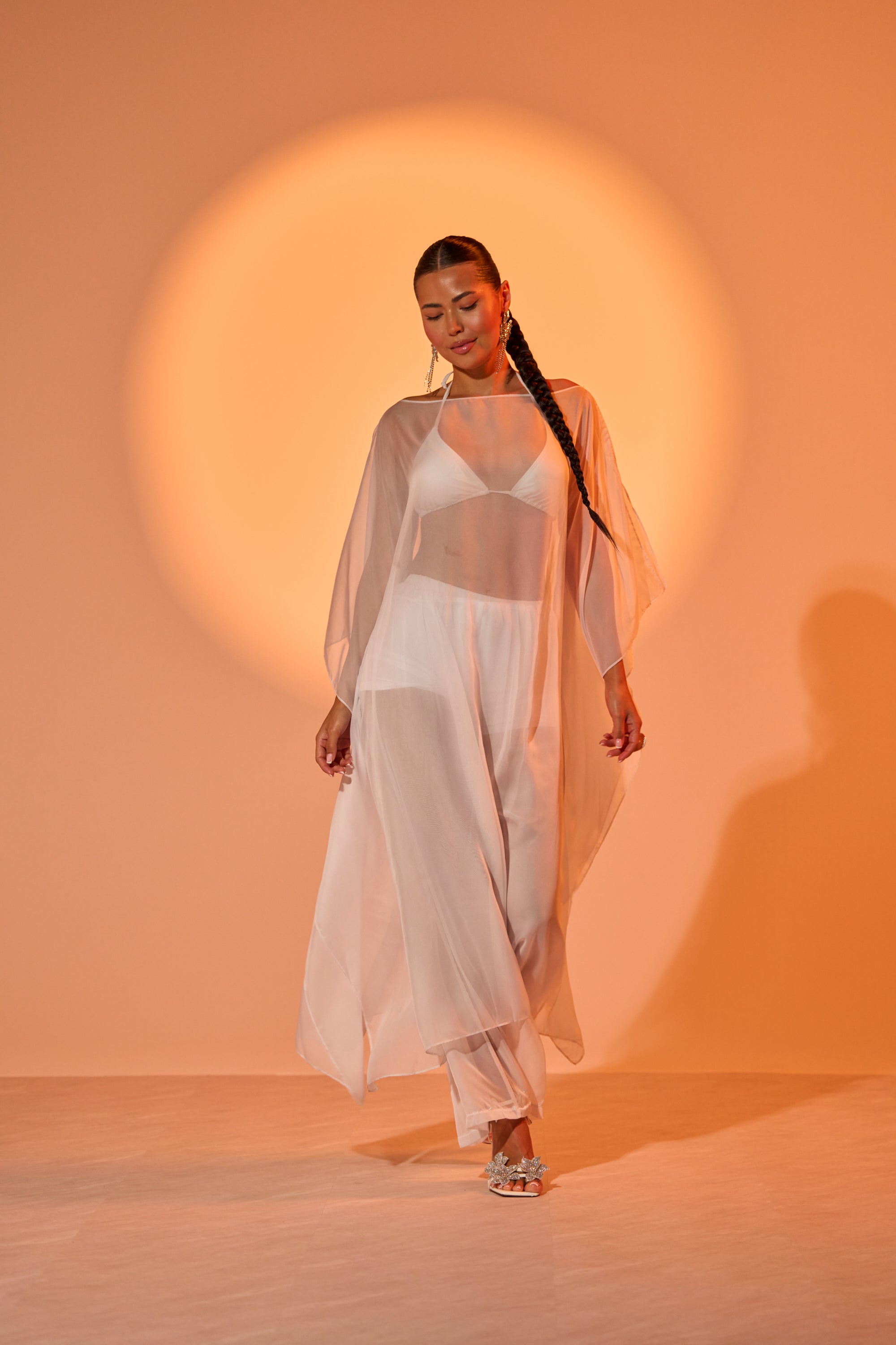 A woman with a long braid walks confidently in the DREAMY SHEER BLOUSE and flowing white pants, set against a warm, orange-lit background with a circular spotlight behind her. 1
