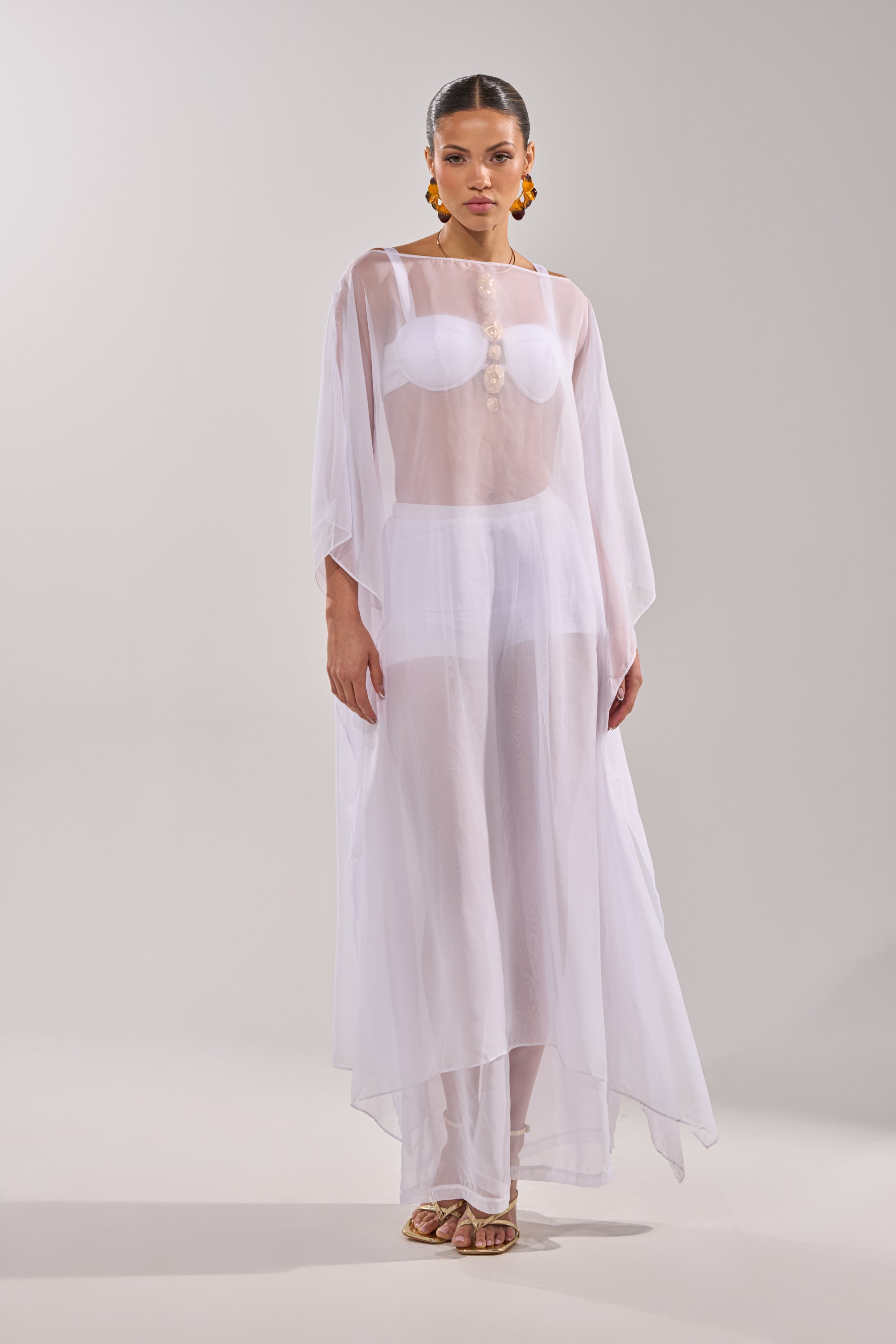 A woman poses against a light gray background, wearing the DREAMY SHEER BLOUSE styled as a flowing white dress over a bikini set. She accessorizes with slicked-back hair, large orange earrings, and gold sandals. 5