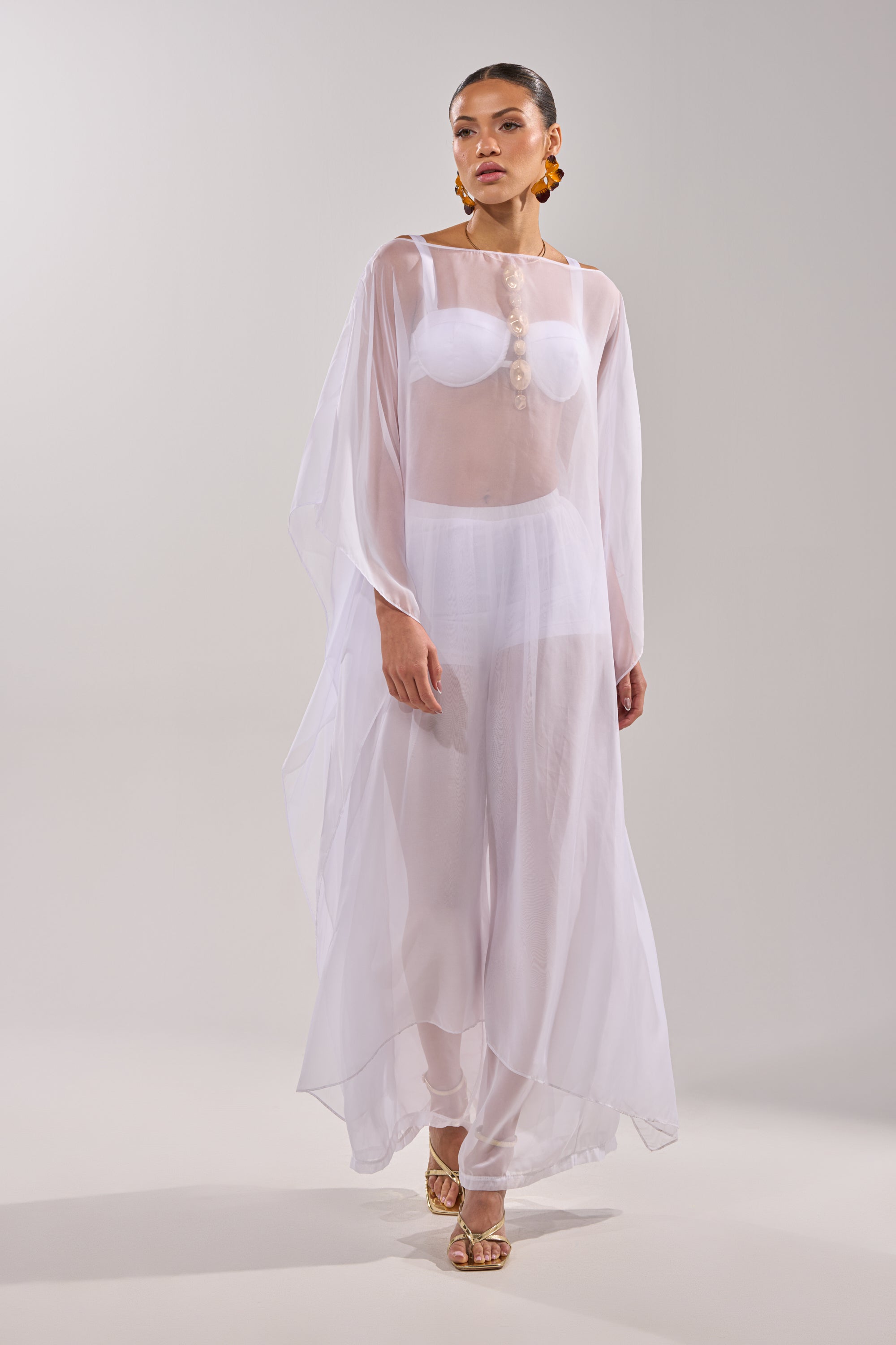 A woman models the DREAMY SHEER BLOUSE over a white bra and briefs, styled with statement earrings and gold sandals, posing confidently against a plain light background. 6