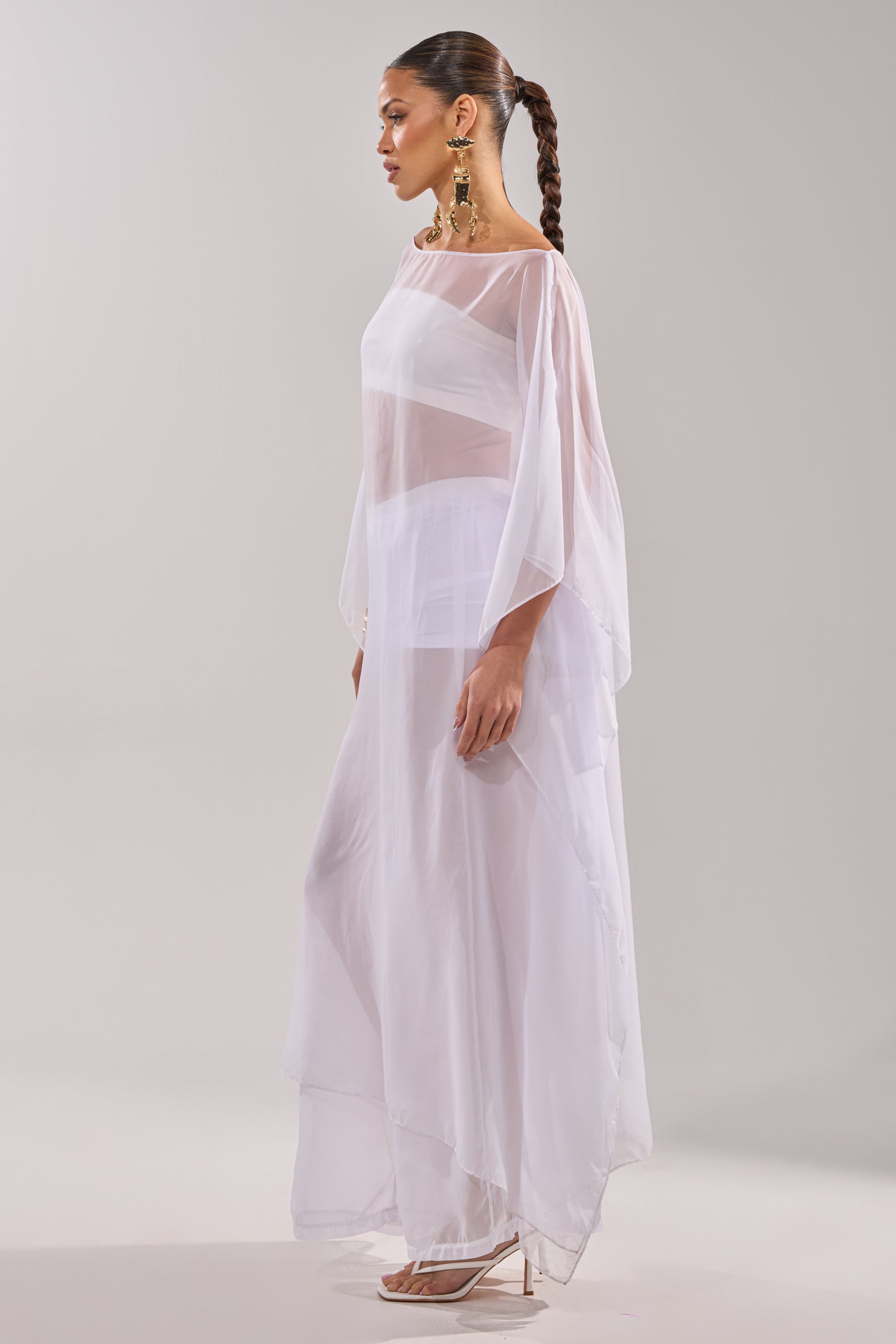 A woman poses in profile against a plain background, wearing the DREAMY SHEER BLOUSE with asymmetrical layers and long sleeves. She accessorizes with gold statement earrings, white high-heeled sandals, and a sleek braided ponytail. 7