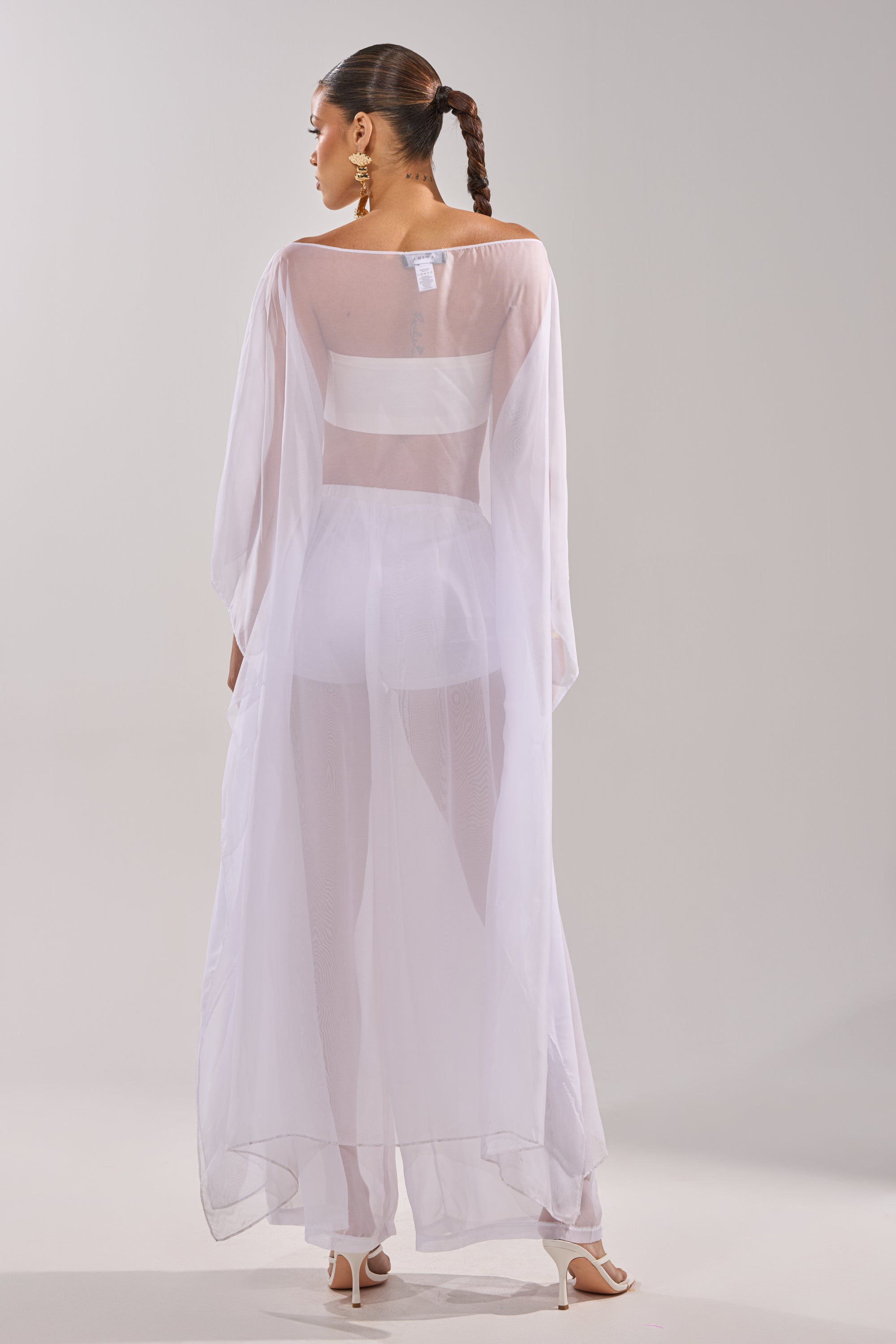 A woman faces away in the DREAMY SHEER BLOUSE, paired with flowing white pants and heels. Her hair is styled in a long braid and she wears gold statement earrings, set against a simple light background. 8