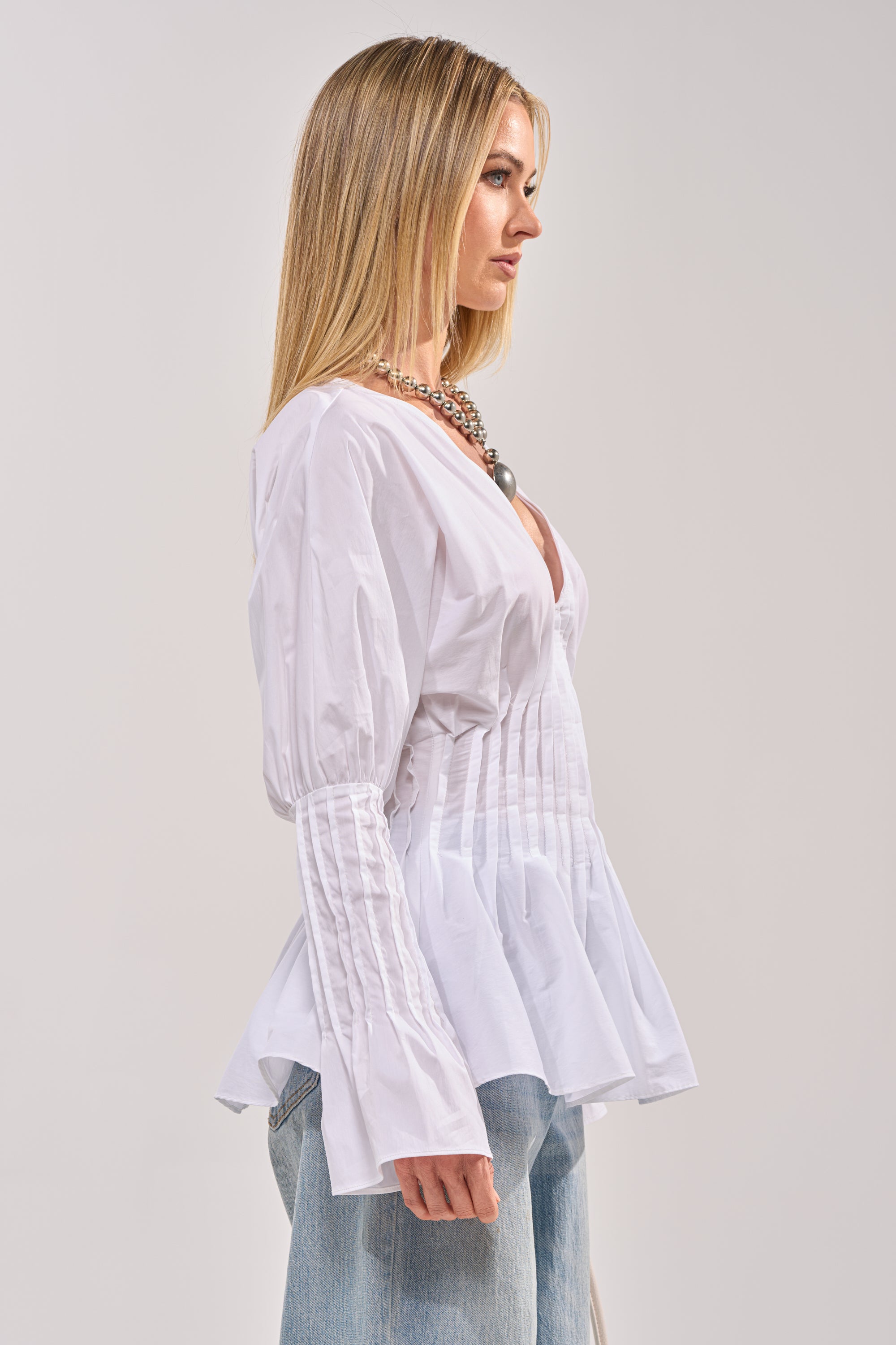A woman with long blonde hair wears the DONT LOOK AT ME BLOUSE, a pleated white long-sleeve top, paired with light blue jeans. She stands in profile against a plain light background. 2