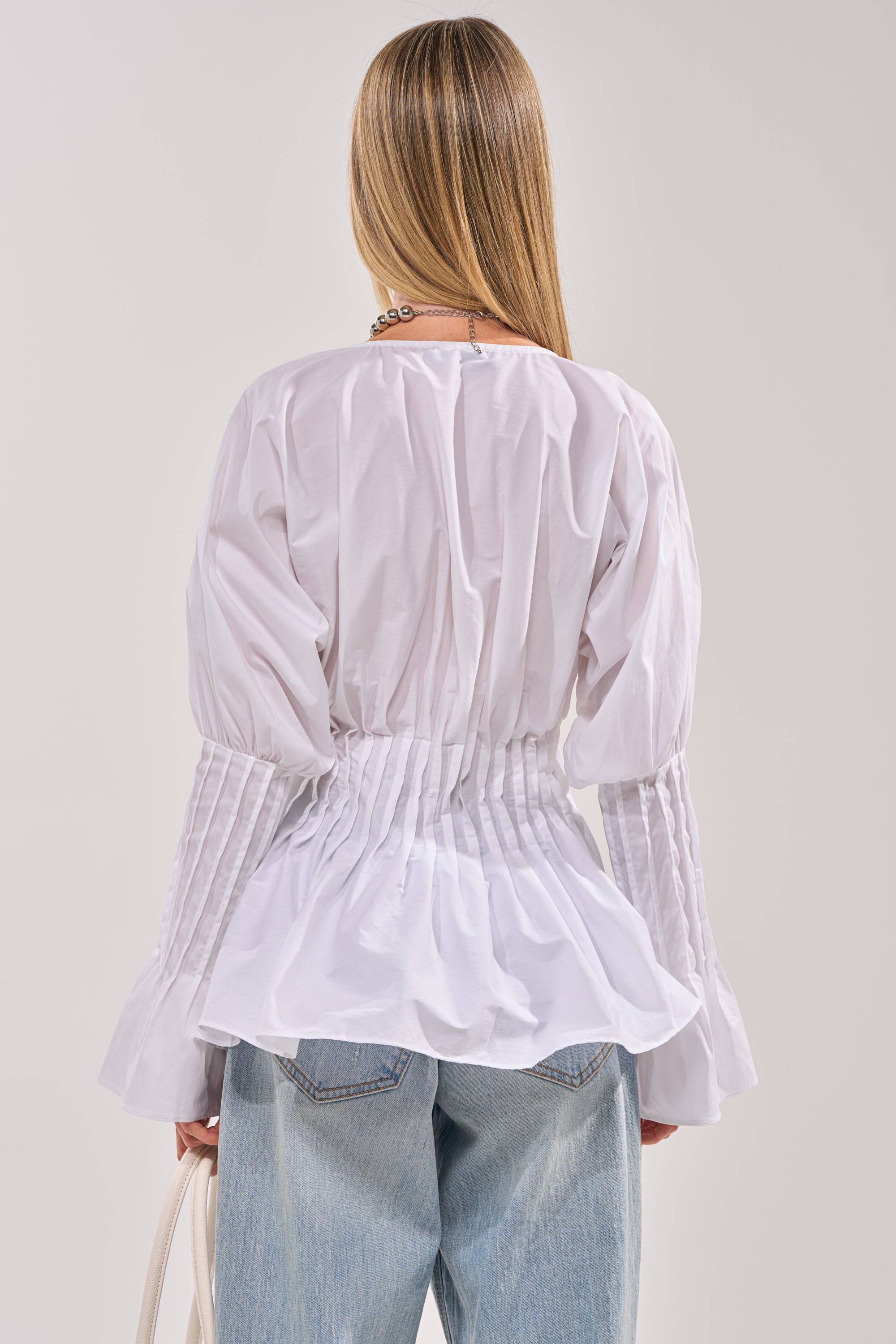 A woman with straight blonde hair, seen from behind, wears the DONT LOOK AT ME BLOUSE—a white, cinched-waist balloon sleeve top—with blue jeans. She holds a white bag and wears a gold necklace. 3