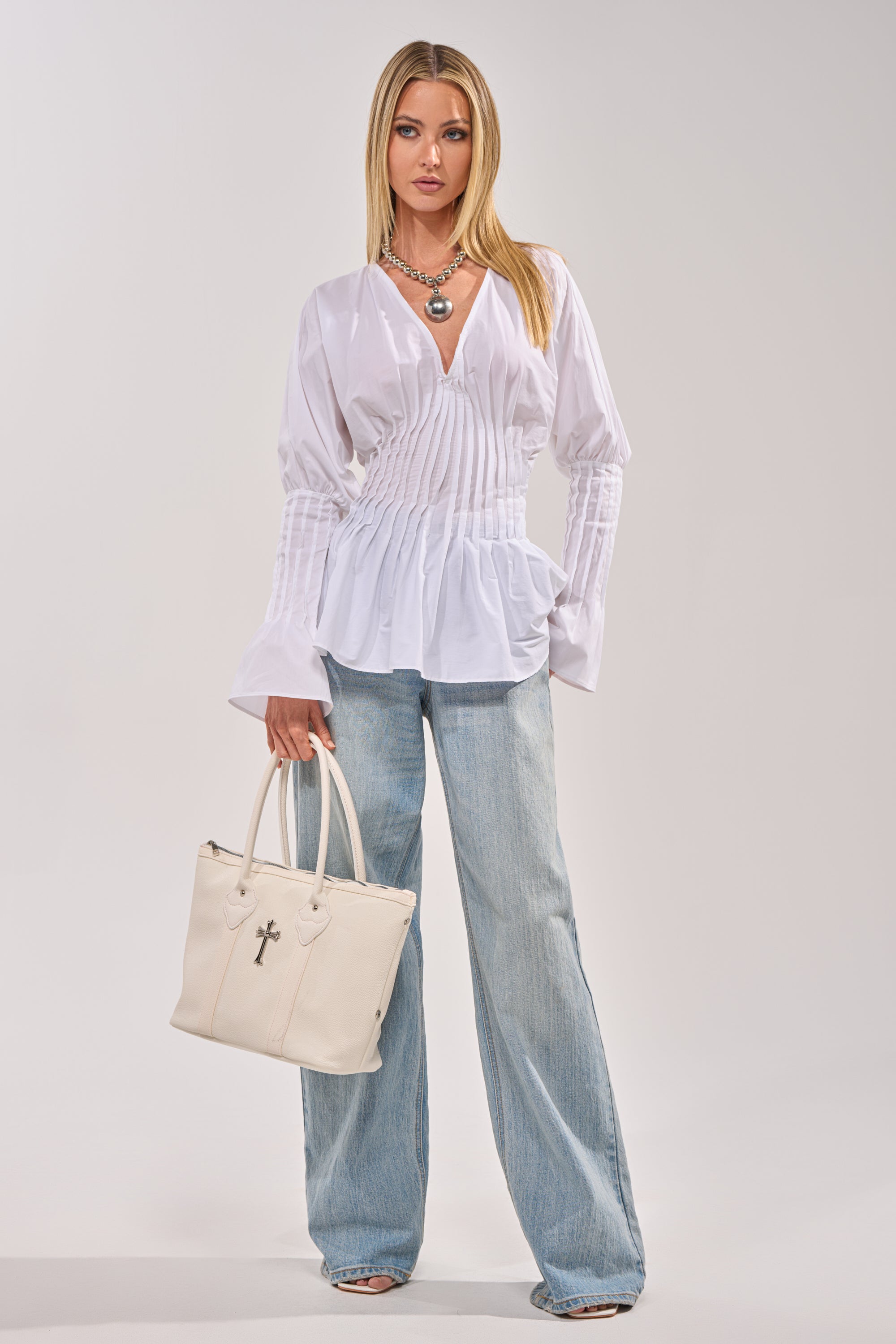 A woman stands against a plain background wearing the DONT LOOK AT ME BLOUSE, a pleated white poplin top with long sleeves, paired with light blue wide-leg jeans, layered silver necklaces, and holding a large white handbag. 4
