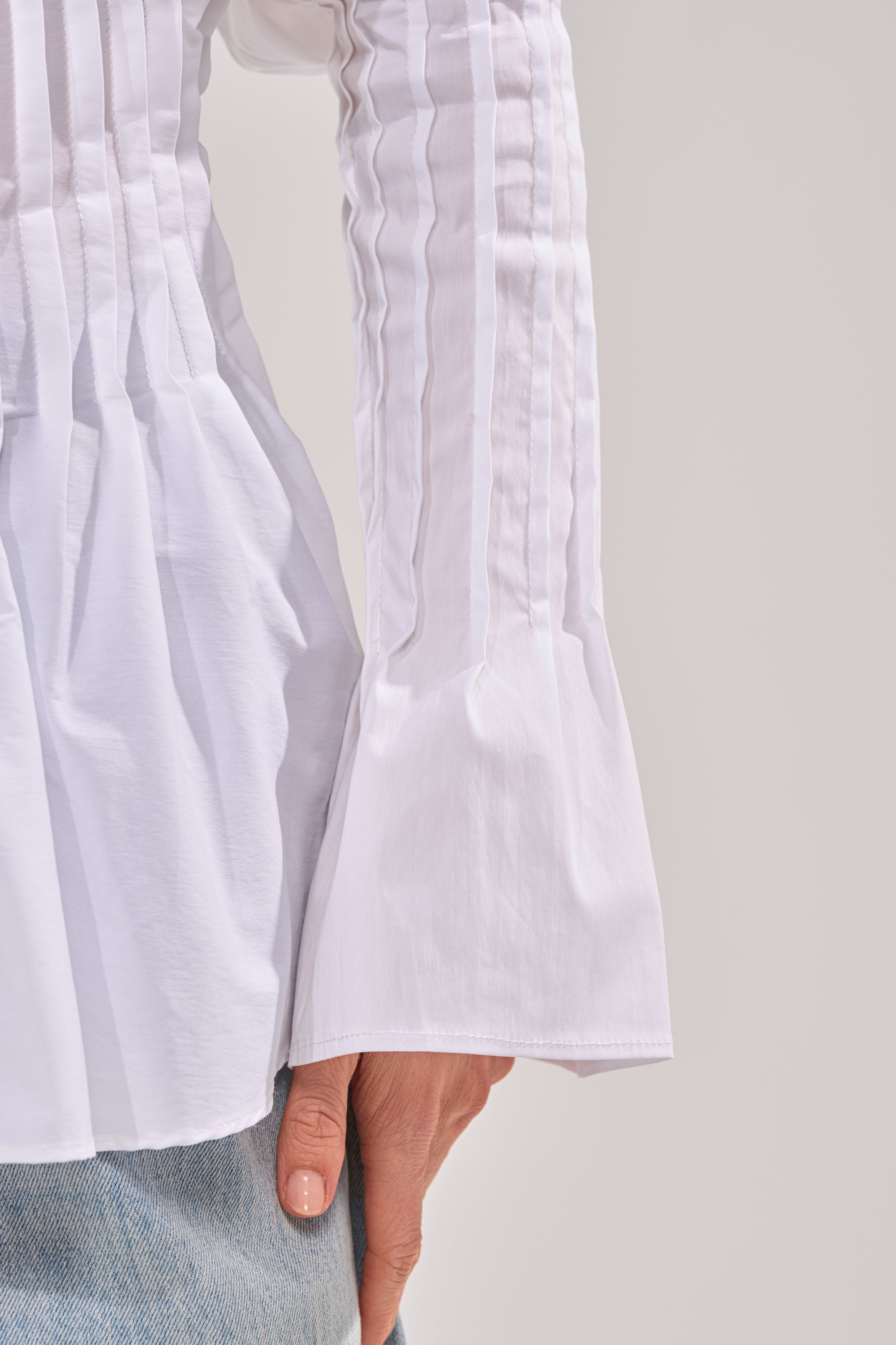 A close-up of a person wearing the DONT LOOK AT ME BLOUSE—a pleated white top with flared cuffs—paired with light blue jeans, hand resting on their hip. 6