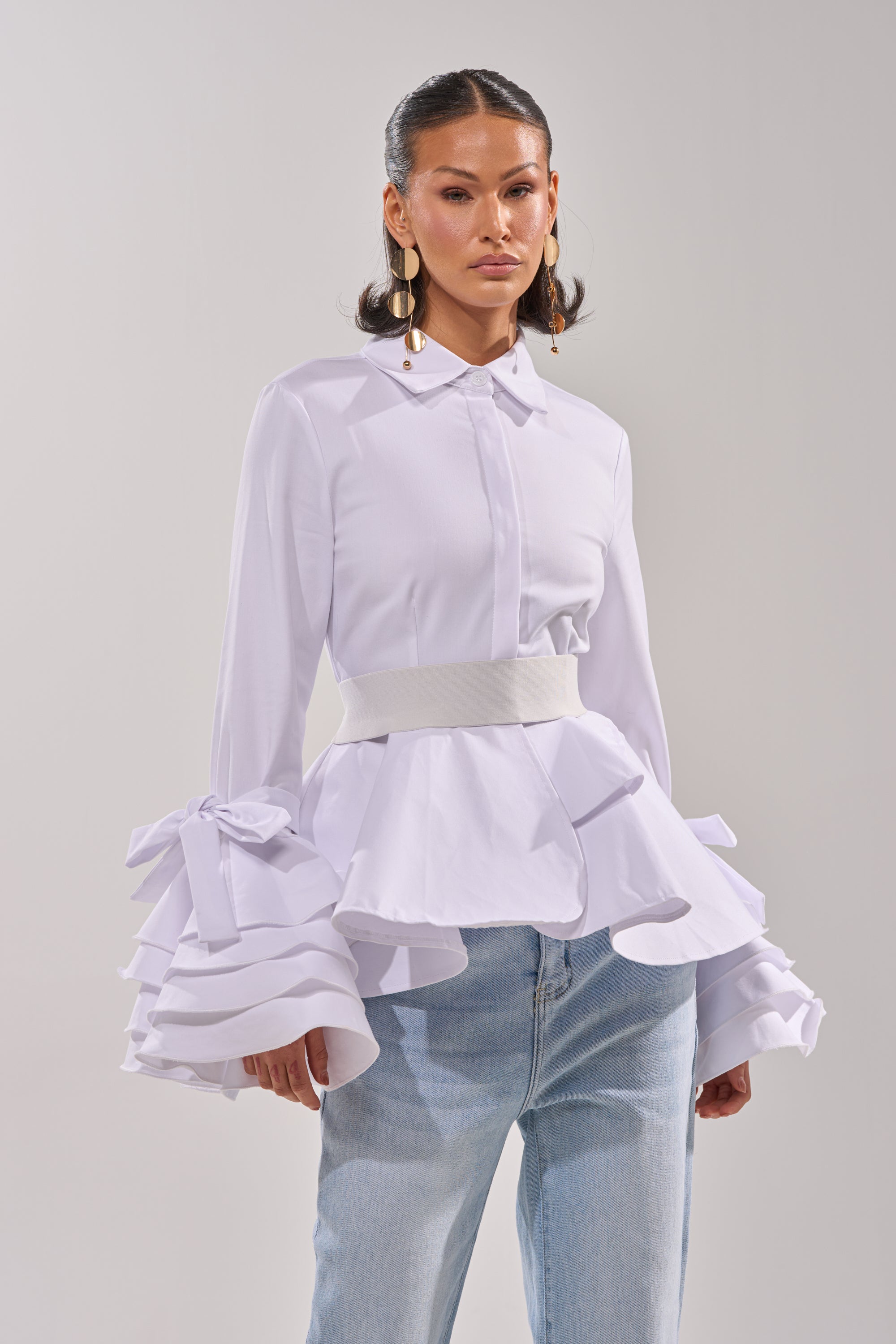A woman models the ELEGANT PEPLUM BUTTON DOWN BLOUSE featuring tiered ruffle cuffs and a wide waistband, paired with light blue jeans. She accessorizes with long earrings, hair styled back, against a plain light background. 1