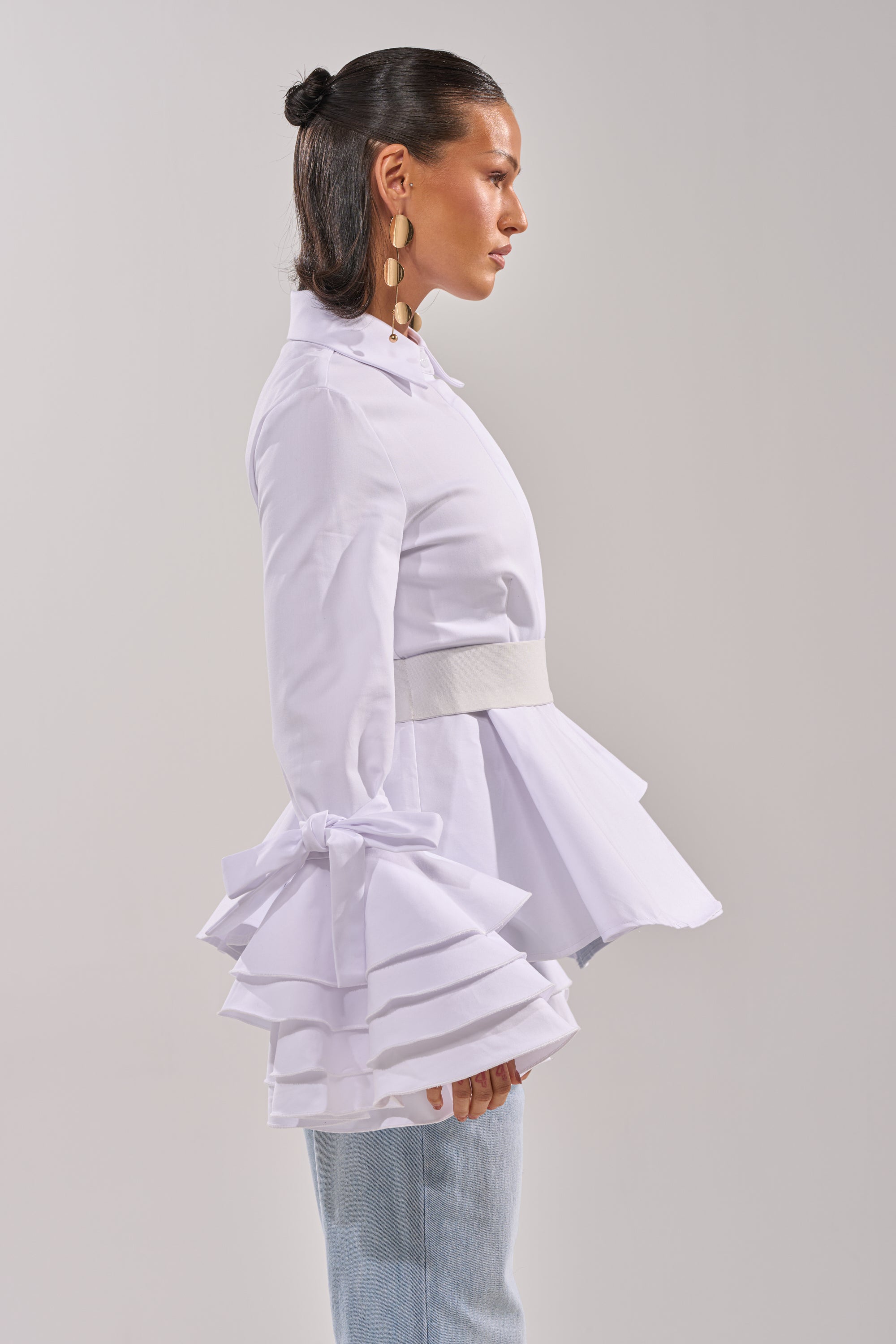 A woman in profile wears the ELEGANT PEPLUM BUTTON DOWN BLOUSE with tiered ruffle cuffs tied with bows, paired with light blue jeans. Her hair is slicked back in a bun, accessorized with large geometric gold earrings. 2