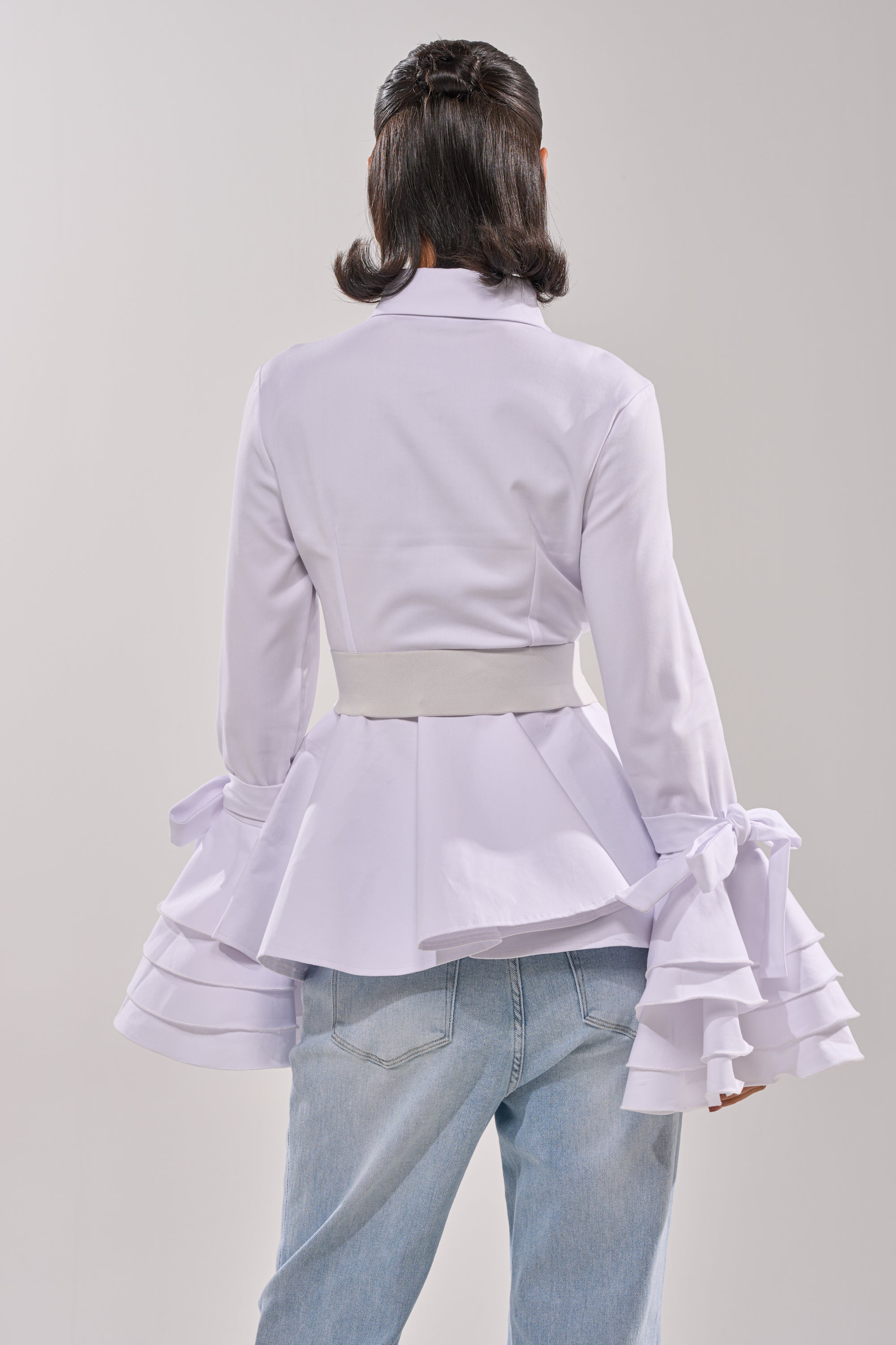A person stands facing away in the ELEGANT PEPLUM BUTTON DOWN BLOUSE with tiered ruffle cuffs and a wide belt, paired with light blue jeans. Their neat, shoulder-length bob is styled against a plain, light background. 3