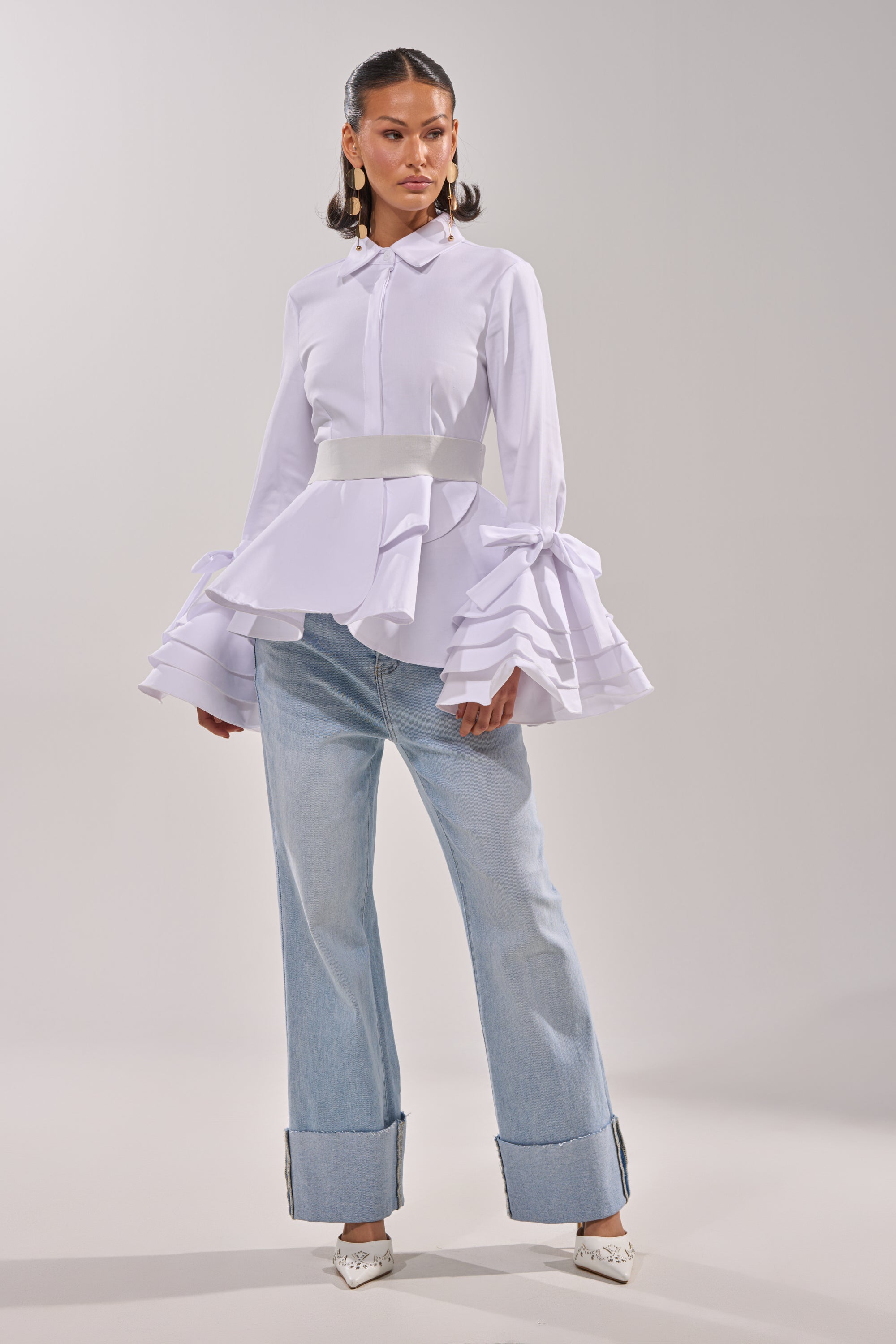 A woman poses in a studio wearing the ELEGANT PEPLUM BUTTON DOWN BLOUSE with tiered ruffle cuffs and a wide belt, styled with light blue wide-leg cuffed jeans, pointed heels, and statement earrings. 4