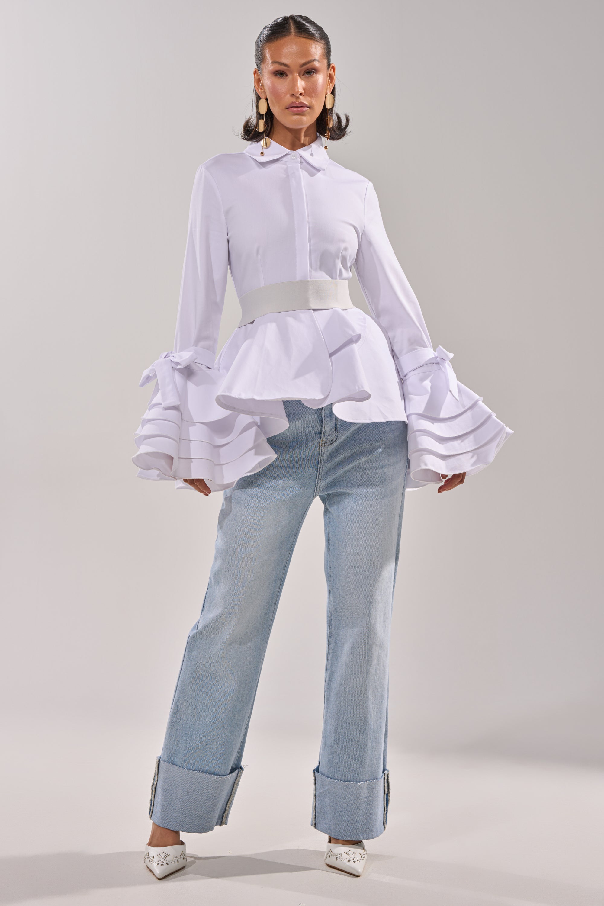 A woman poses confidently in the ELEGANT PEPLUM BUTTON DOWN BLOUSE with tiered ruffle cuffs, styled with light blue cuffed jeans and white pointed heels against a plain light background. 5
