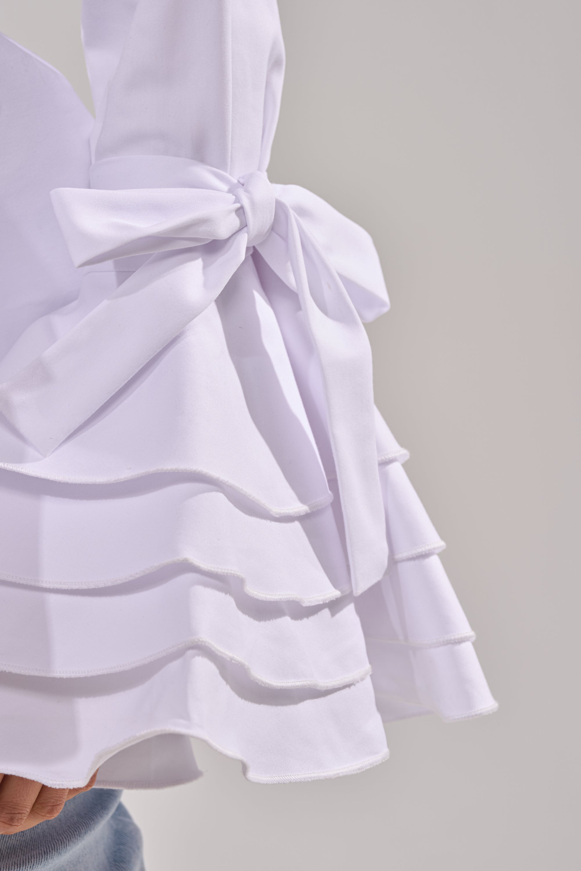 A close-up of the ELEGANT PEPLUM BUTTON DOWN BLOUSE in white, showcasing layered ruffles and a prominent side bow, set against a plain light background. 7