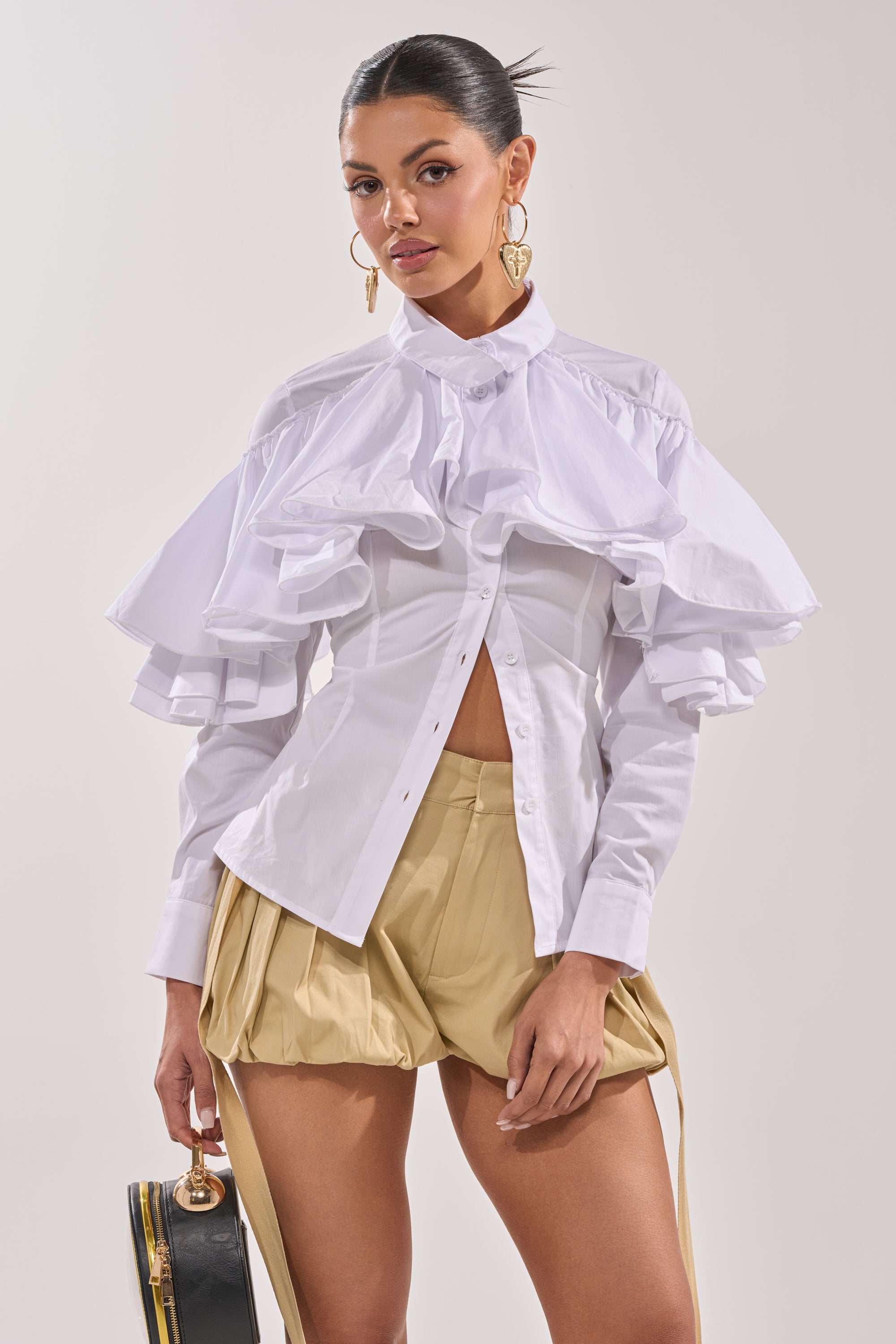 A woman models the CRAZY ENOUGH BUTTON DOWN, a white blouse with ruffle sleeves, paired with khaki pleated shorts with suspenders. She accessorizes with a black handbag and gold hoop earrings, her hair slicked back. 0