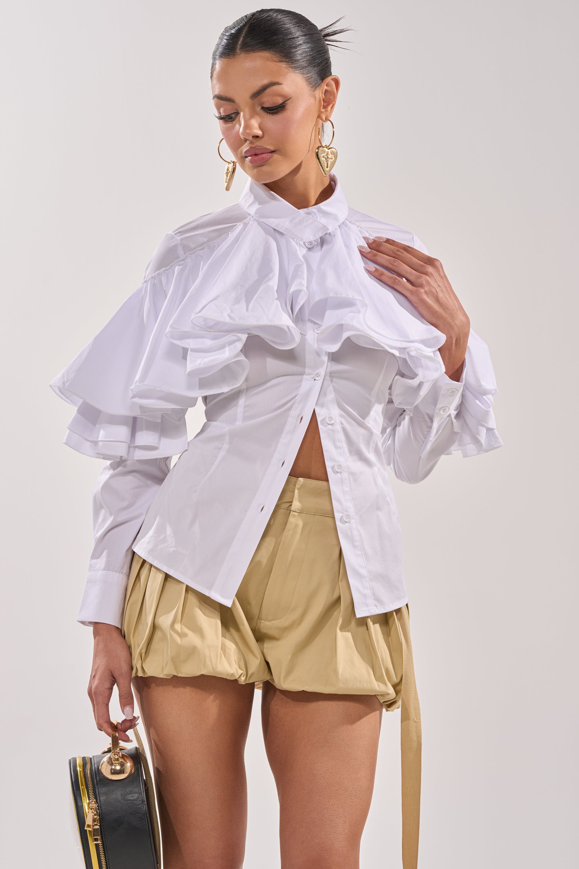 A woman wears the CRAZY ENOUGH BUTTON DOWN—a white ruffle sleeve blouse—and beige pleated shorts, holding a small round black handbag. Her dark hair is tied back and she accessorizes with gold statement earrings. 2