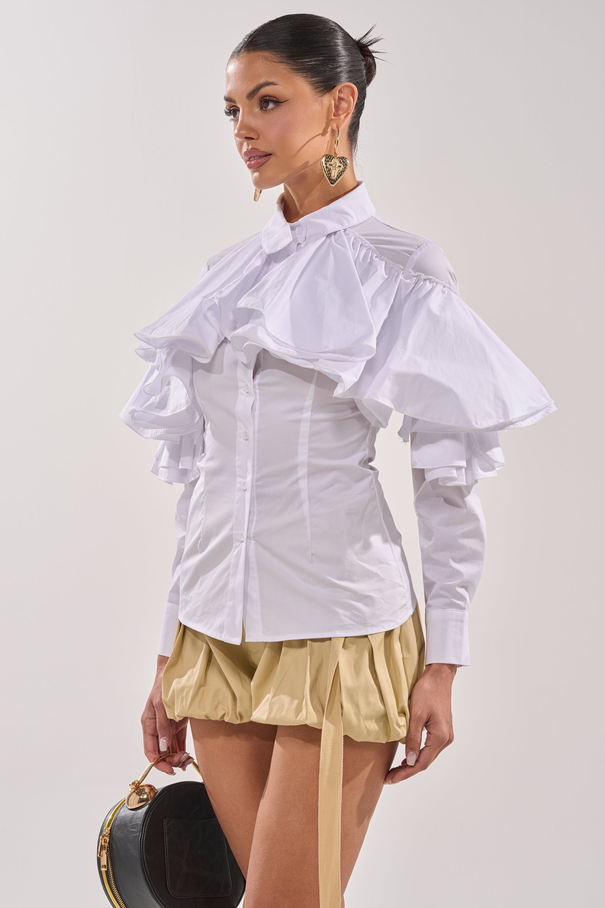 A woman wears the CRAZY ENOUGH BUTTON DOWN, a white blouse with ruffle sleeves, paired with a short pleated beige skirt. Her hair is tied back, she accessorizes with gold earrings and holds a black circular handbag against a plain background. 5