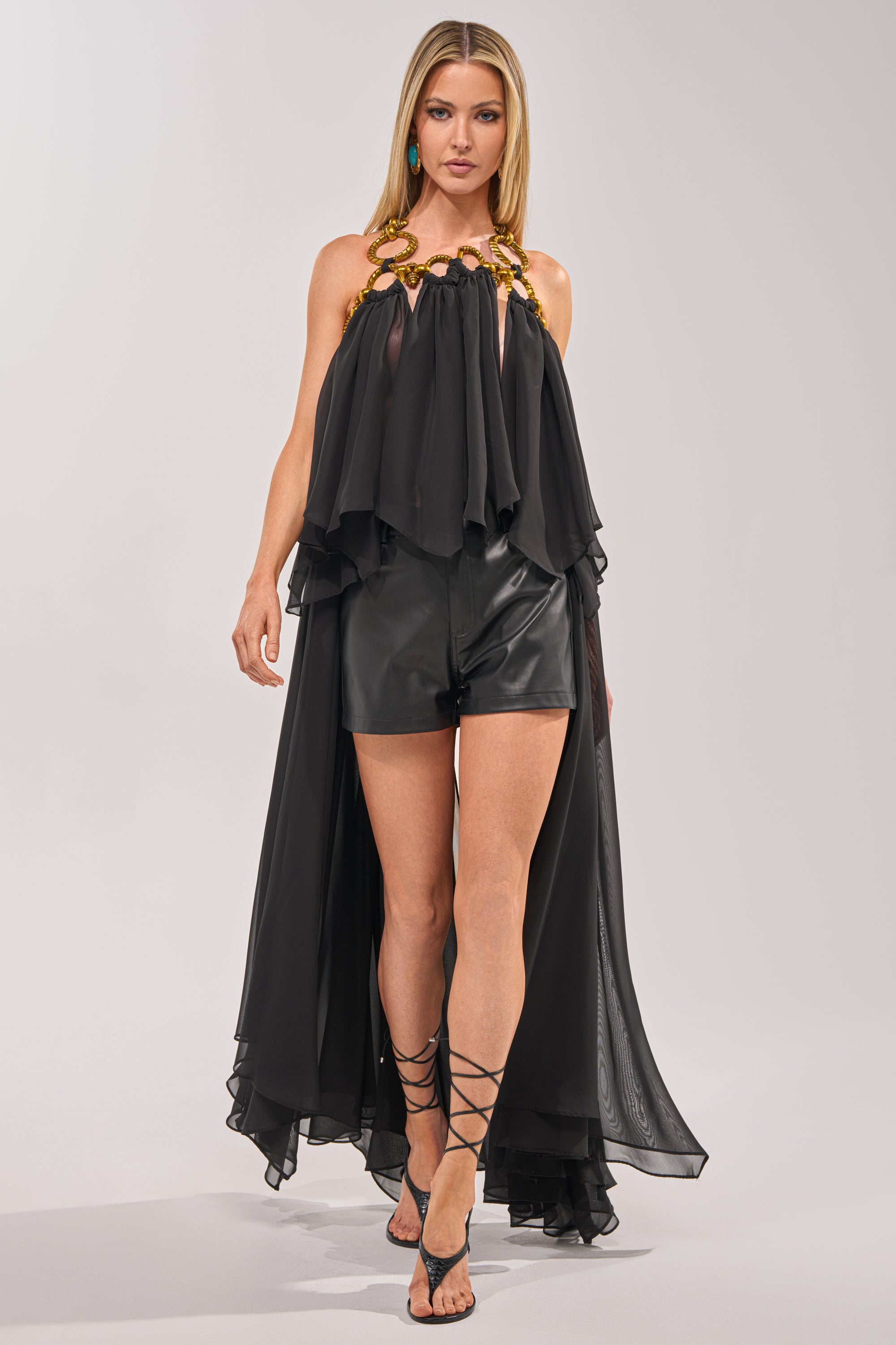 A woman models the DANCE THE NIGHT AWAY BLOUSE—a black organza top with a high-low silhouette and gold chain neckline—paired with black leather shorts and strappy heels for chic evening style against a plain background. 0