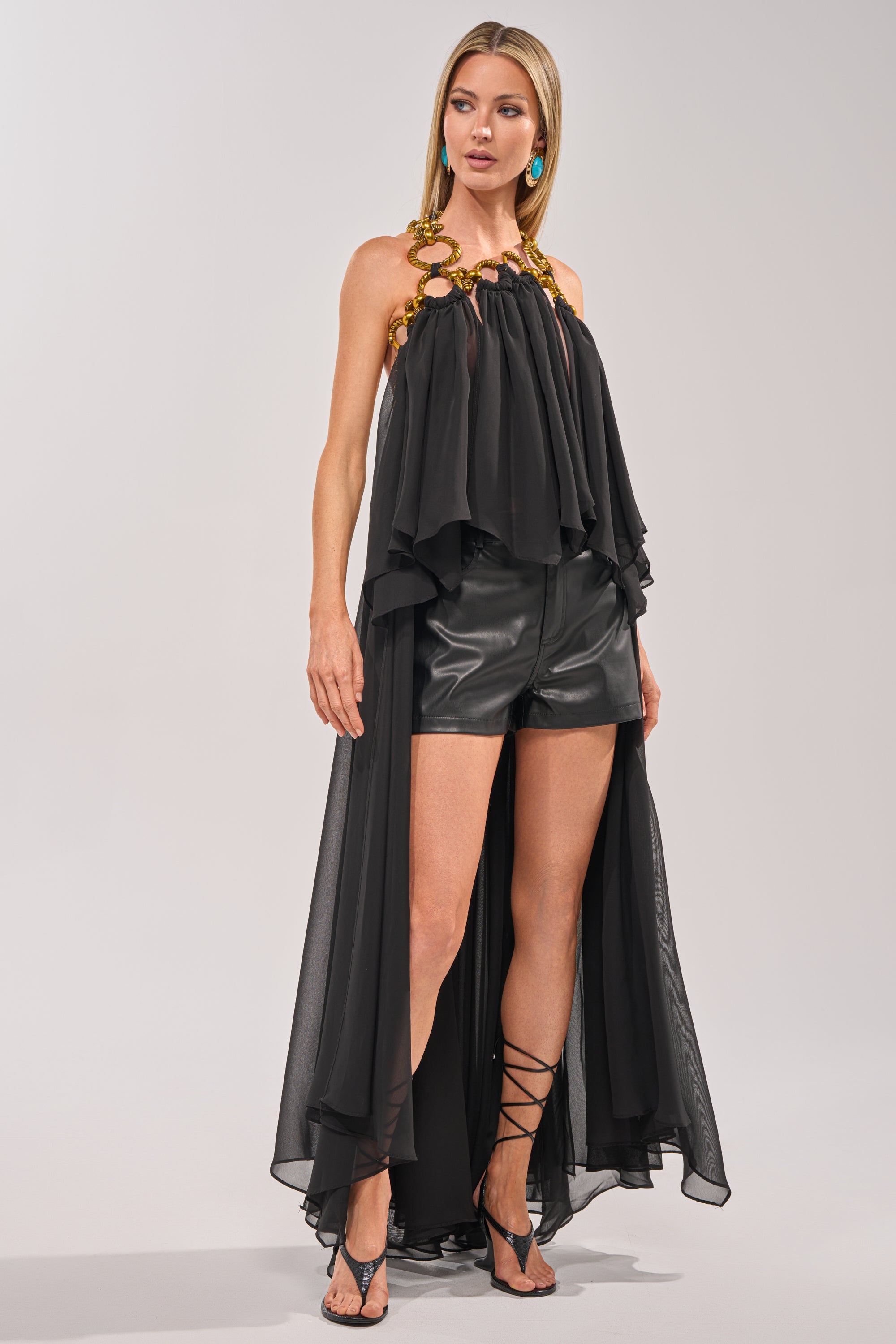 A woman styles the DANCE THE NIGHT AWAY BLOUSE, a black organza high-low halter top with a dramatic long train, over black leather shorts, strappy heels, gold chain accents, and turquoise earrings for a bold evening look against a light background. 1