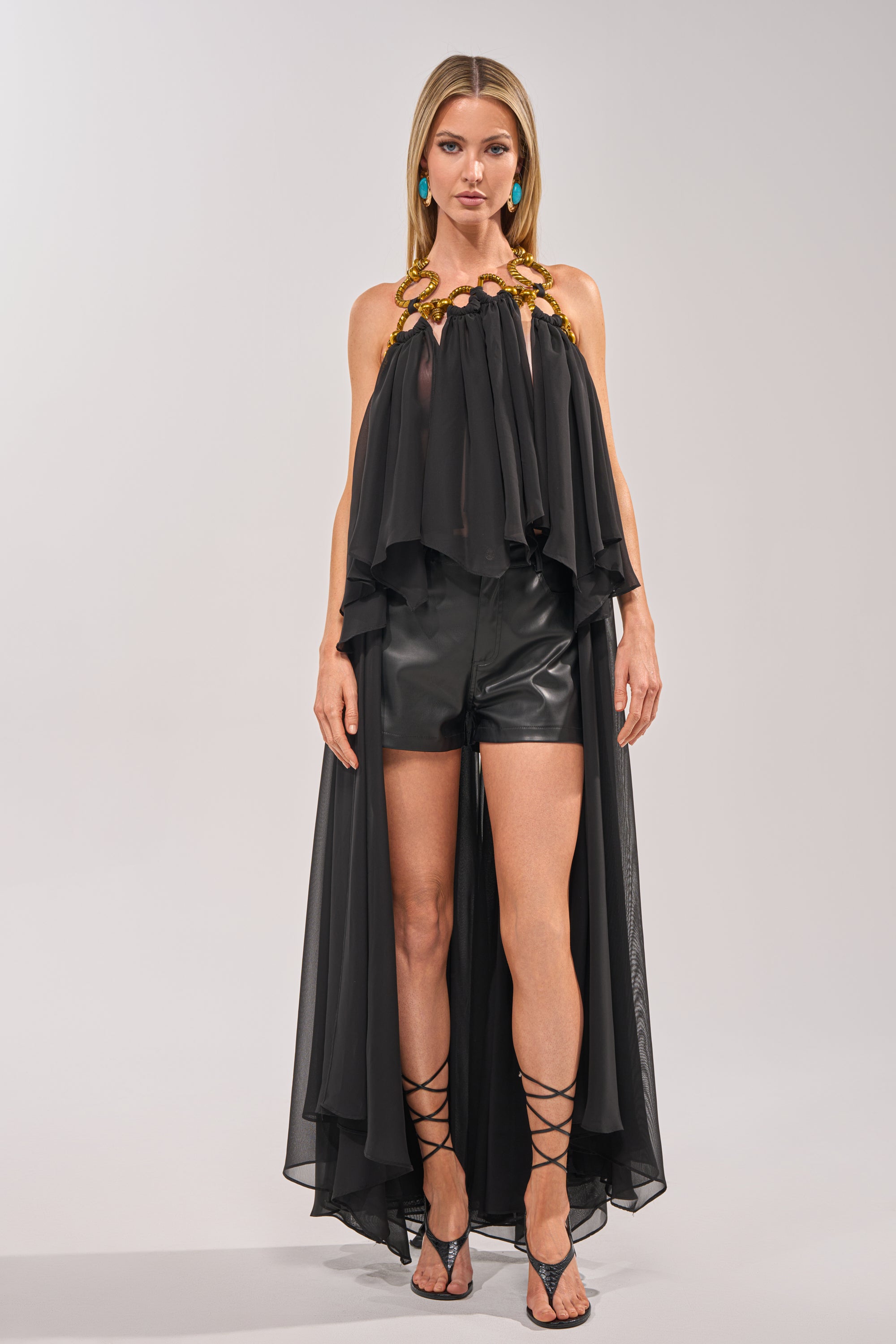 A woman with straight hair and turquoise earrings poses against a plain light background, wearing the DANCE THE NIGHT AWAY BLOUSE—a black organza high-low halter top with gold accents—paired with black leather shorts and lace-up sandals. 2
