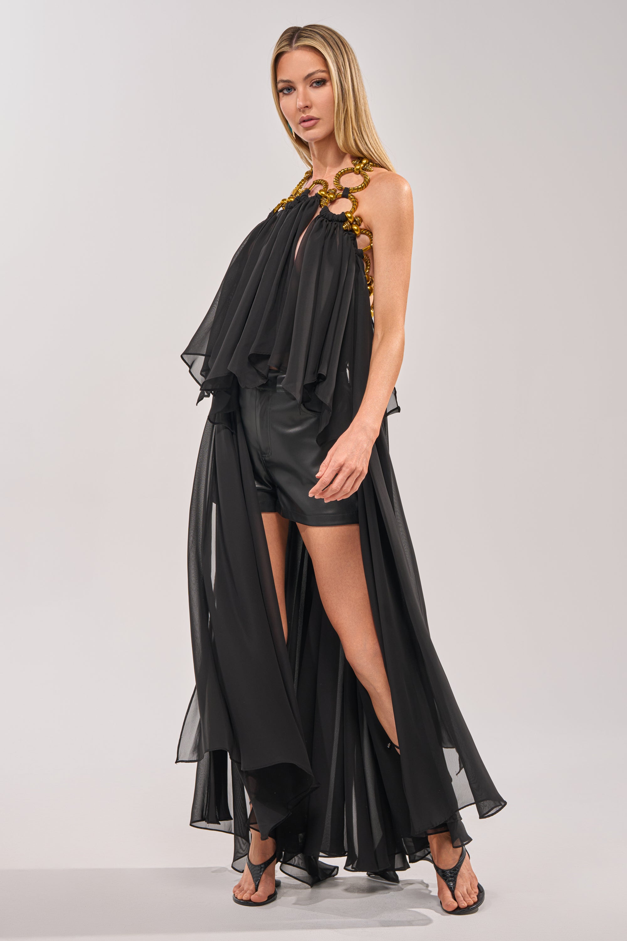 A woman with long blonde hair stands against a light gray background, wearing the DANCE THE NIGHT AWAY BLOUSE—a flowing black top with gold shoulder details, halter neckline, sheer layers over black shorts—and black sandals. 3