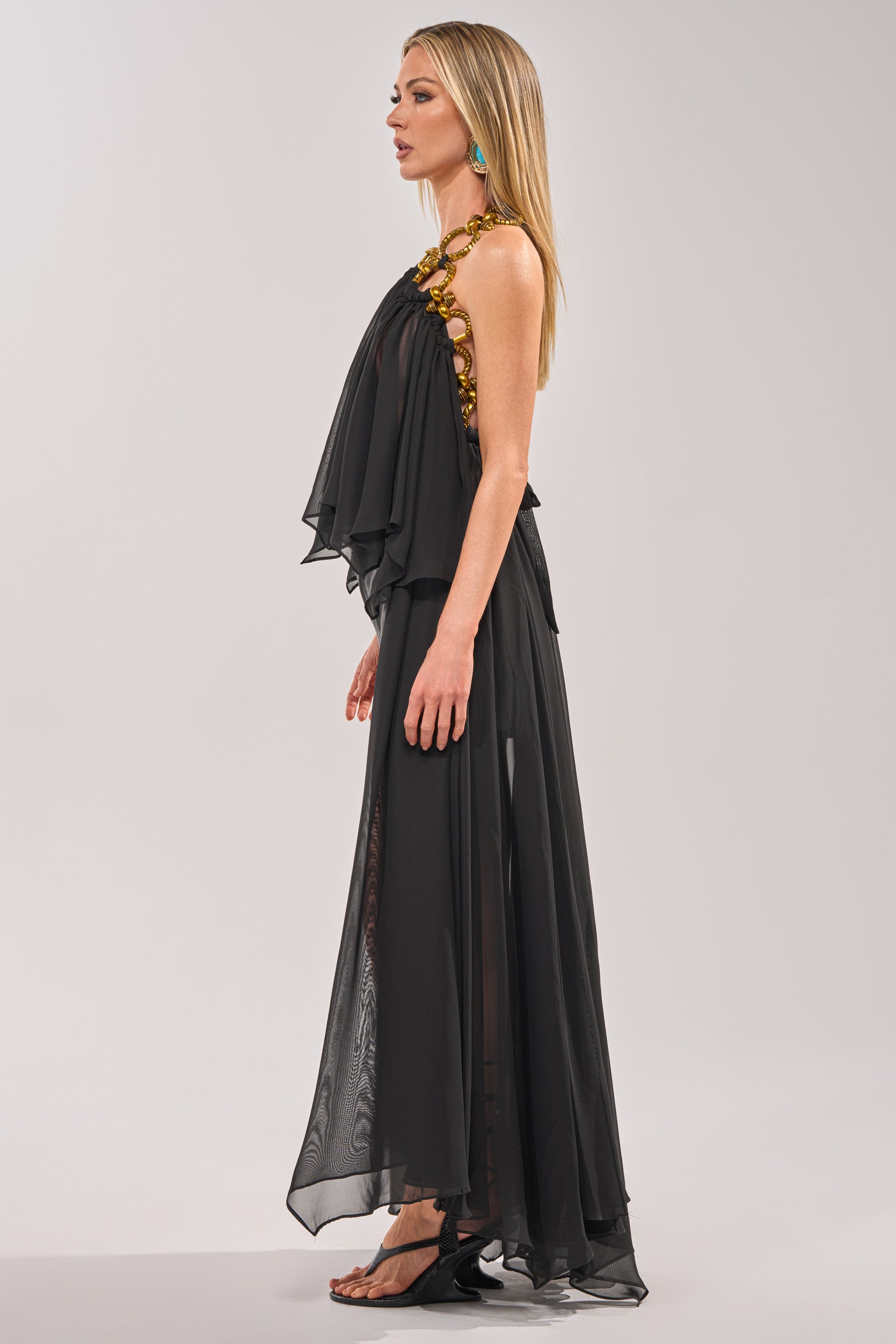 A woman poses in profile wearing the DANCE THE NIGHT AWAY BLOUSE with gold floral detail on one shoulder, paired with black sandals and turquoise earrings—an elegant evening look shown against a simple, light backdrop. 4