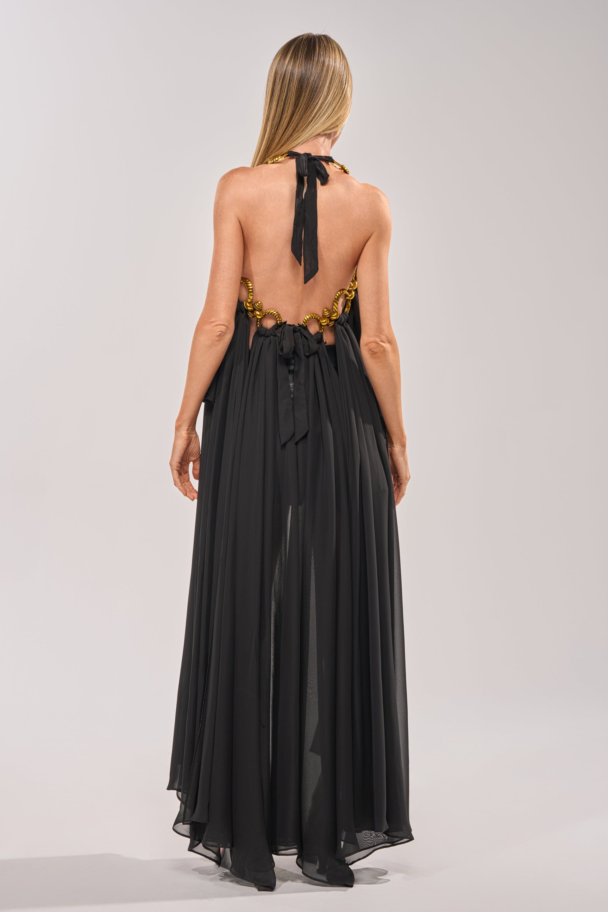 A woman with long blonde hair faces away, wearing the DANCE THE NIGHT AWAY BLOUSE—an elegant black top with a halter neckline, bow tie details, and gold embellishments—perfect for sophisticated evening styling. 5